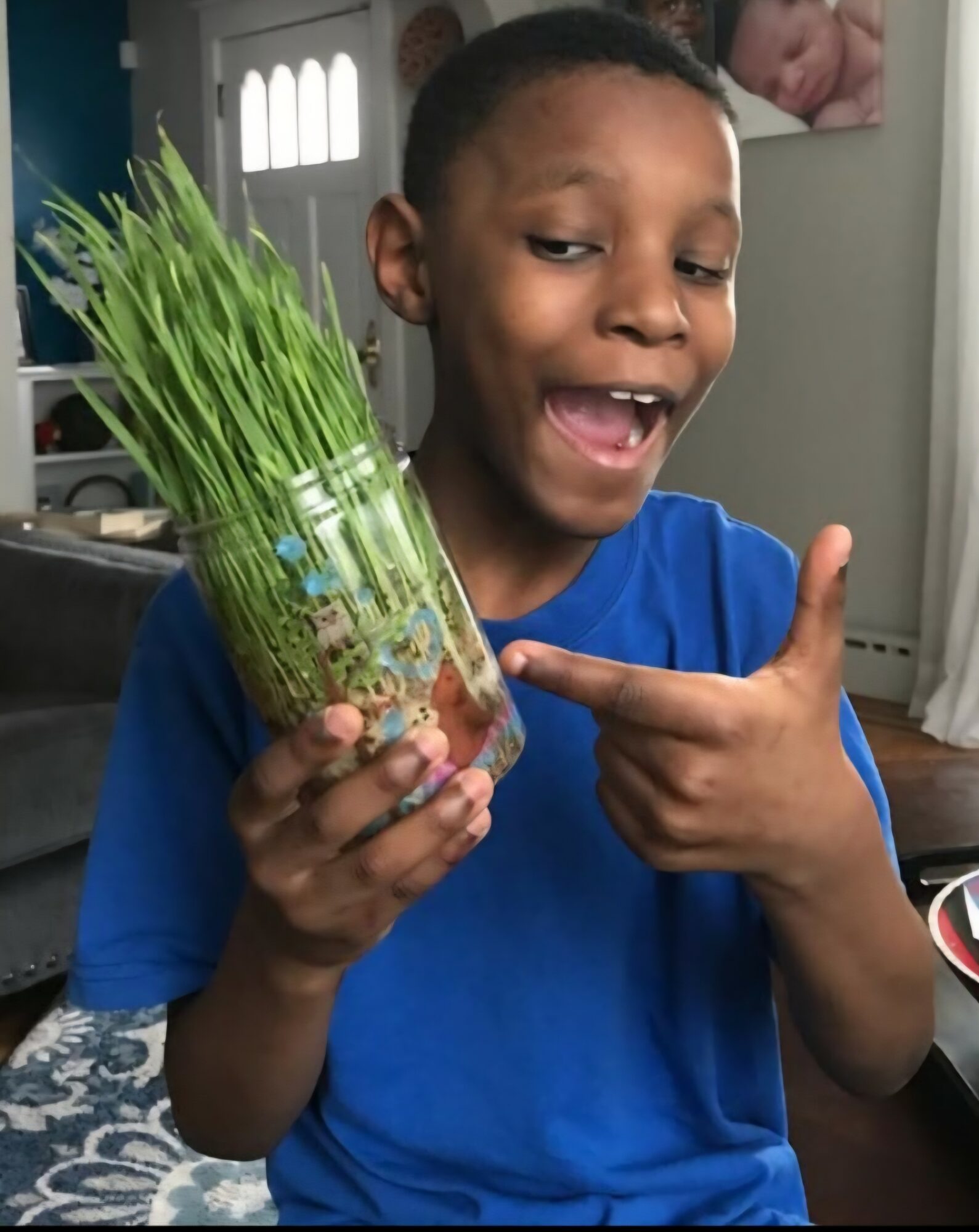 Child holding a glass jar with green grass and smiling, pointing at it indoors.