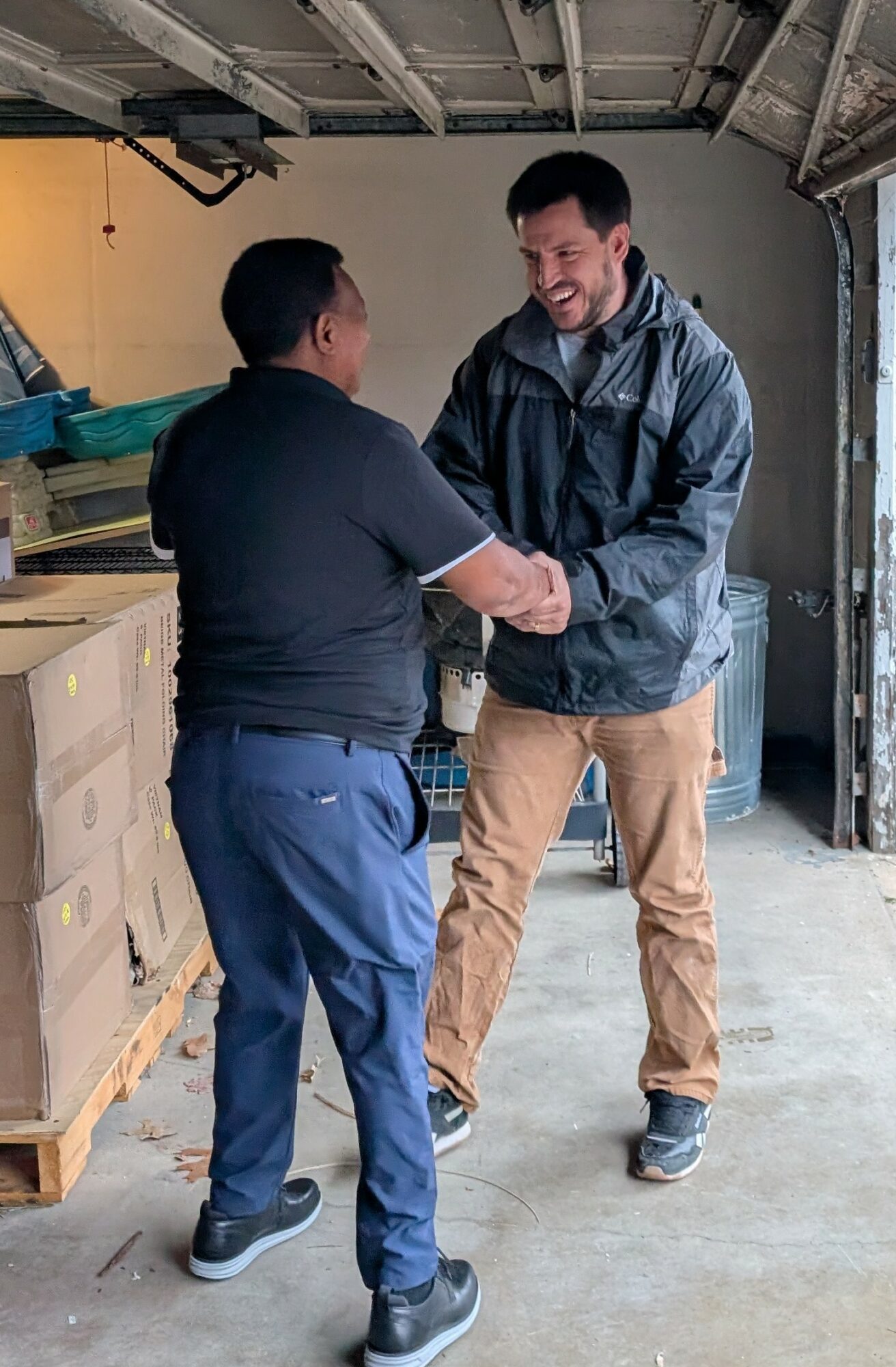 Two men shake hands in a garage or workshop with shelves and tools in background.