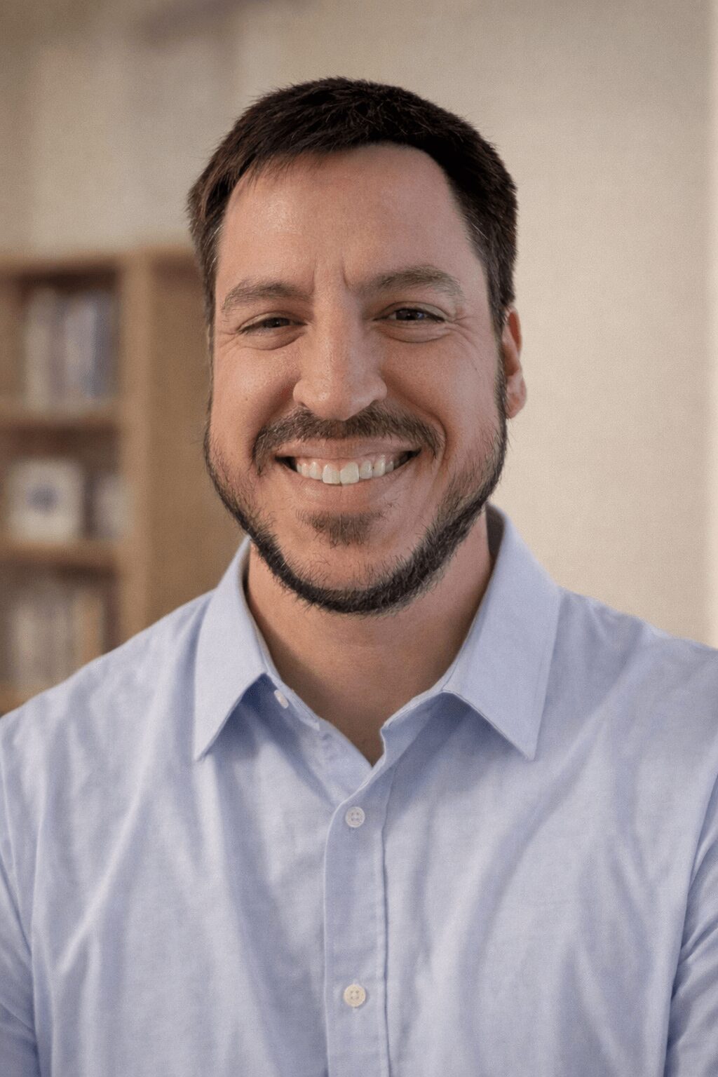 Man with short dark hair and beard smiling, wearing a light blue shirt, in an indoor setting.