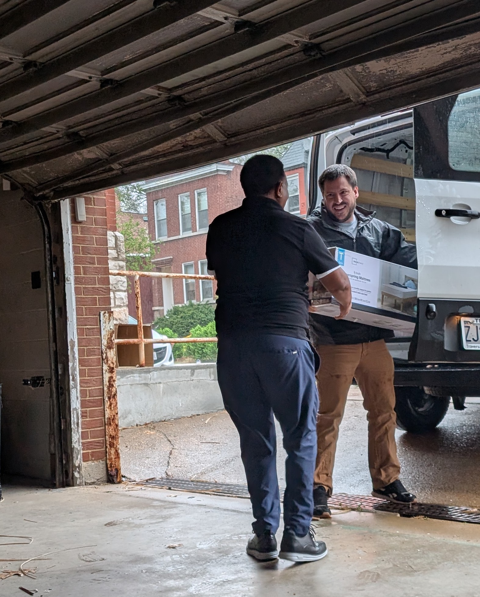 Two men exchanging a package outside a garage, one inside a delivery truck, brick building in background.