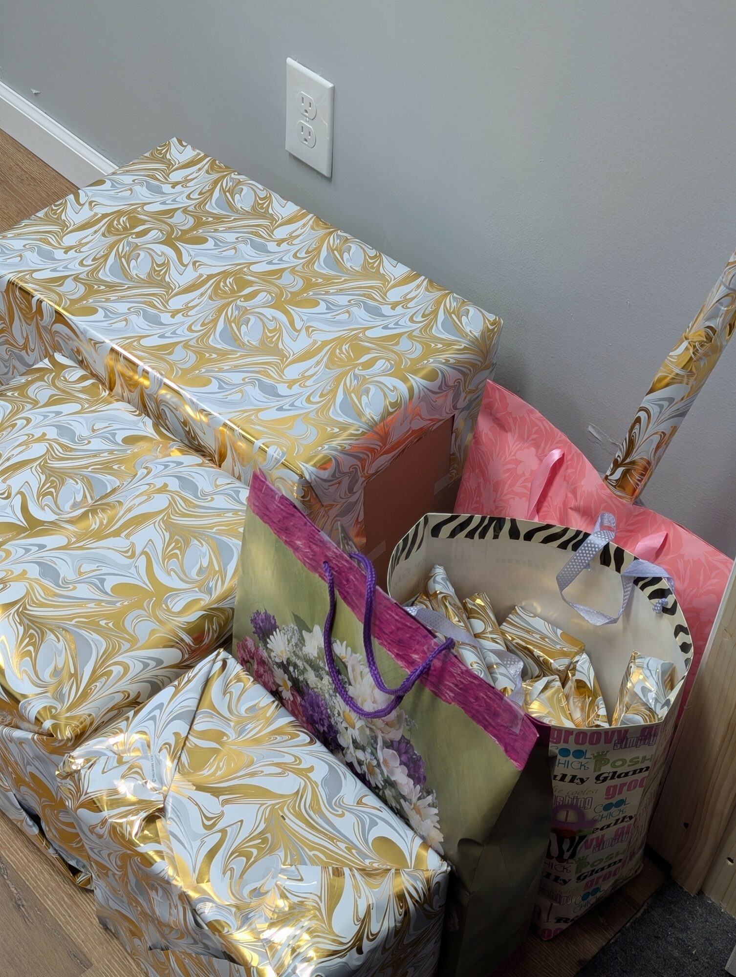 Several wrapped gift boxes and shopping bags stacked near a wall with an electrical outlet.