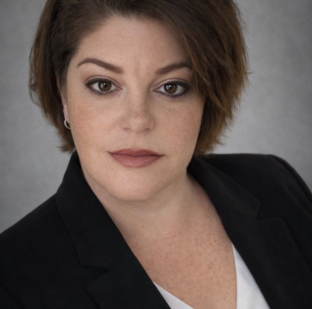 Woman with short brown hair, wearing a black blazer and white top, looking at the camera.