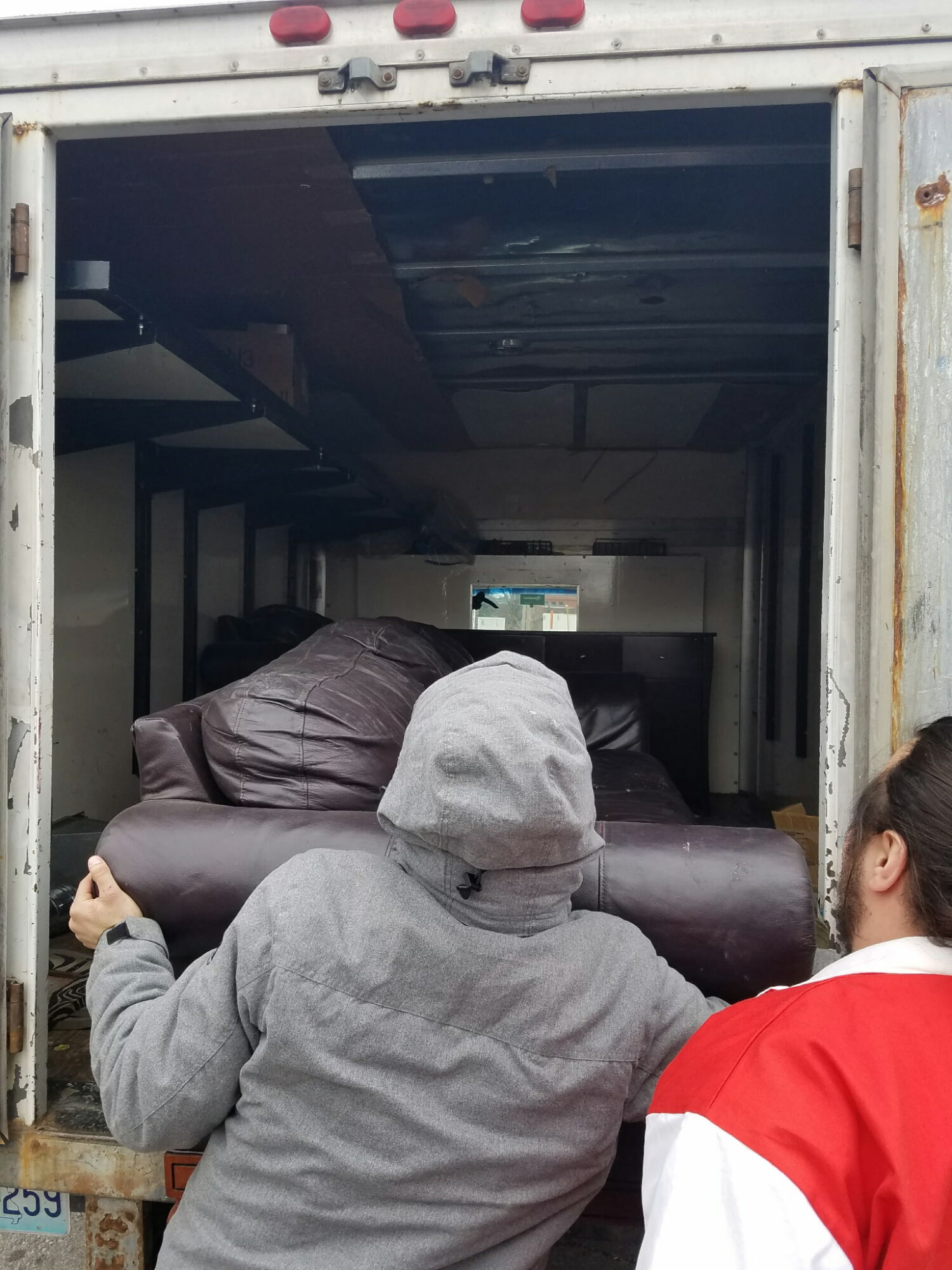 Two people look into a truck with a sofa inside, one wearing a gray hoodie and the other in a red and white jacket.