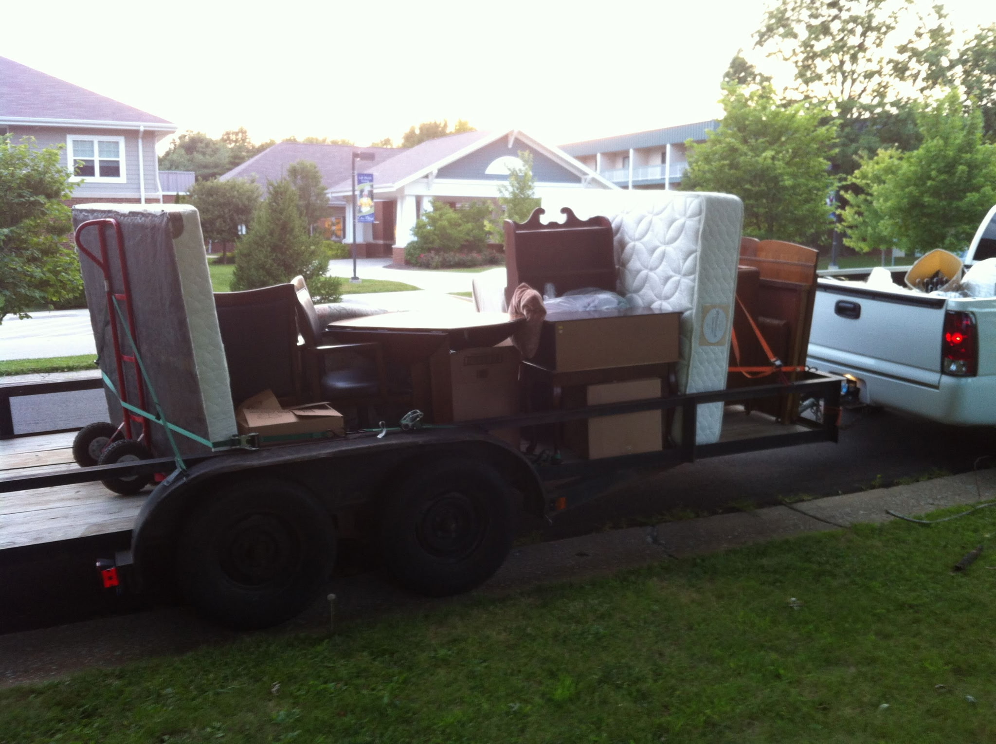 Trailer with various furniture pieces, including a mattress, chairs, and a table, parked on a driveway in a residential neighborhood.