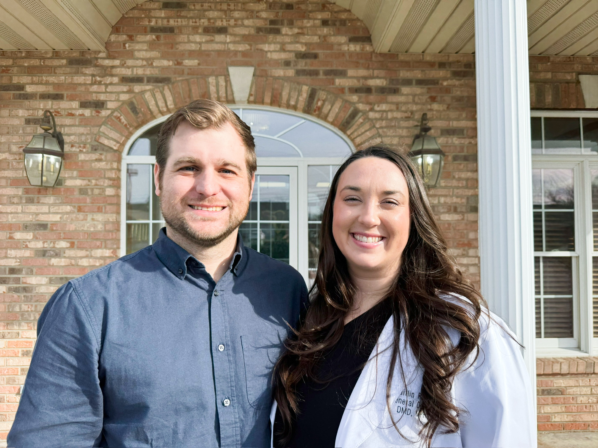 A smiling man and woman stand outside a brick building with large windows and white columns.