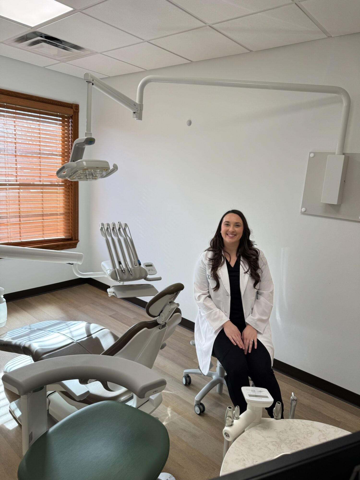 Woman in white coat sitting on dental chair in dental clinic with equipment and window in background.