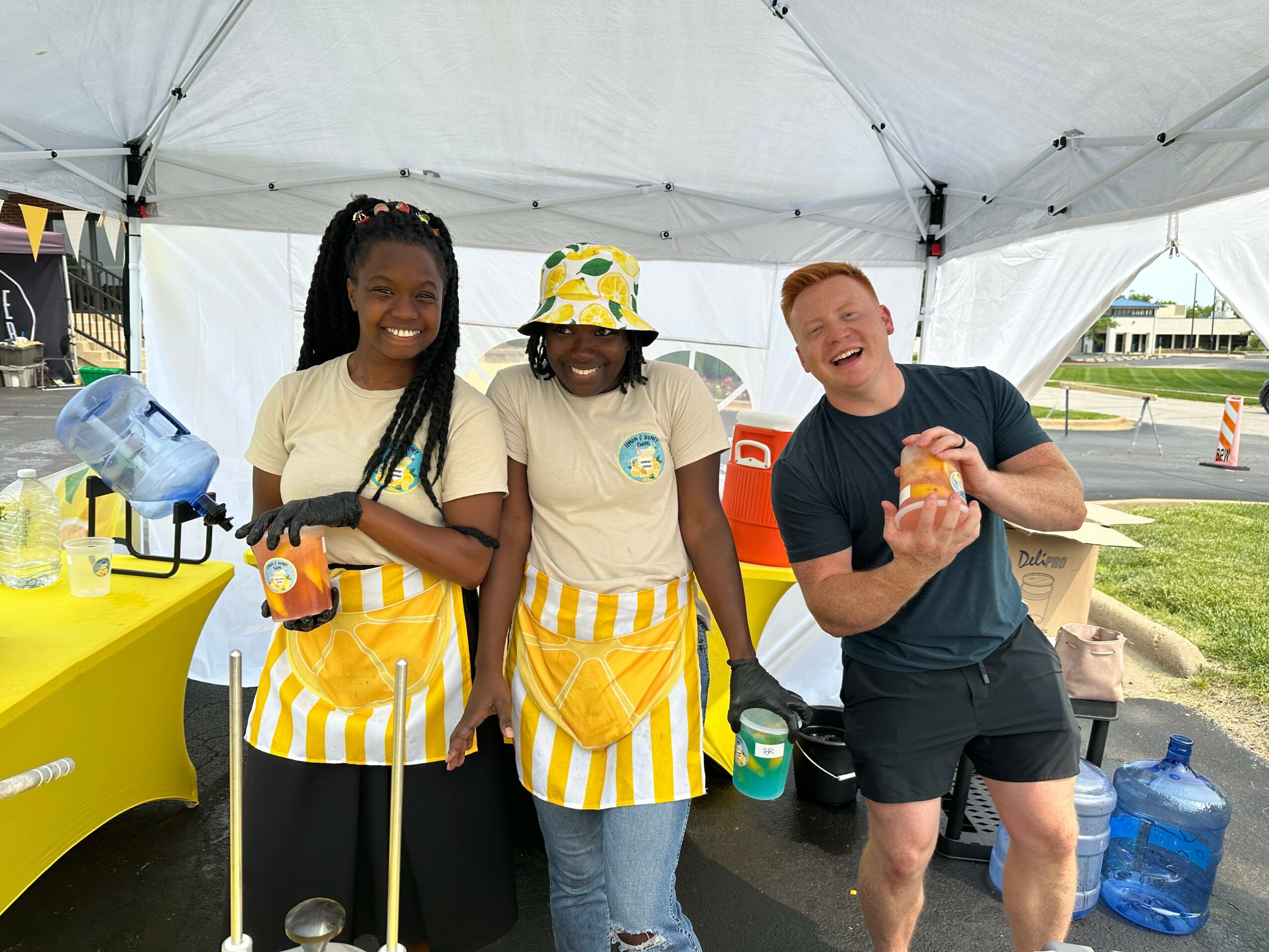 Three people smiling under a tent, two women wearing yellow striped aprons and a man holding a burger.