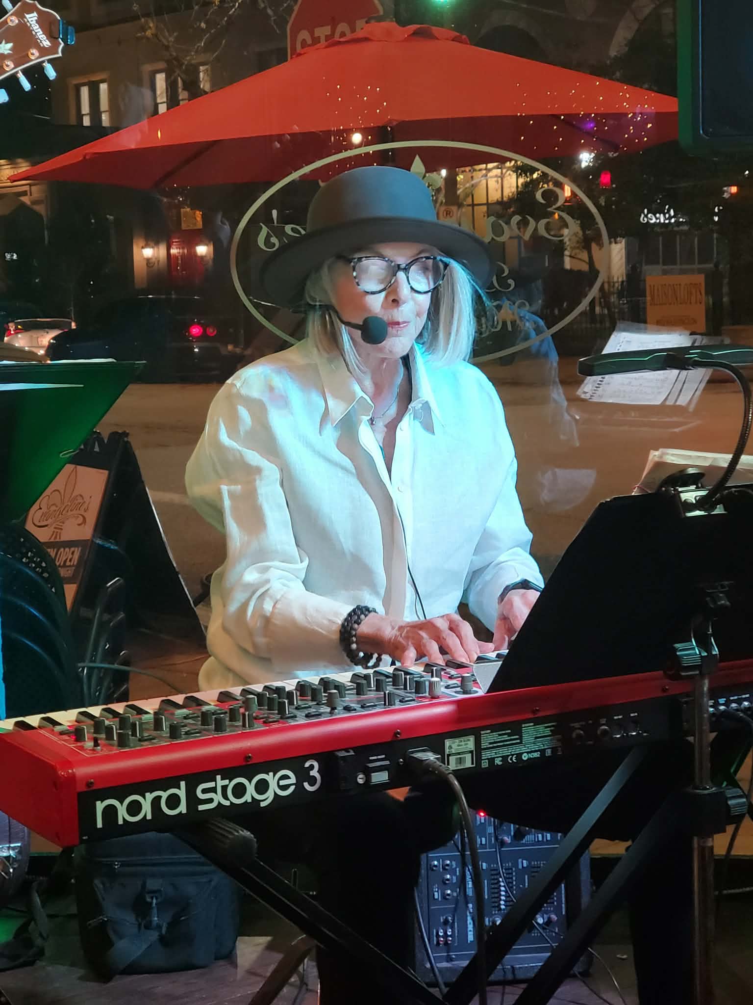 Woman with glasses and a hat playing a keyboard in a cafe at night, with a red umbrella outside.