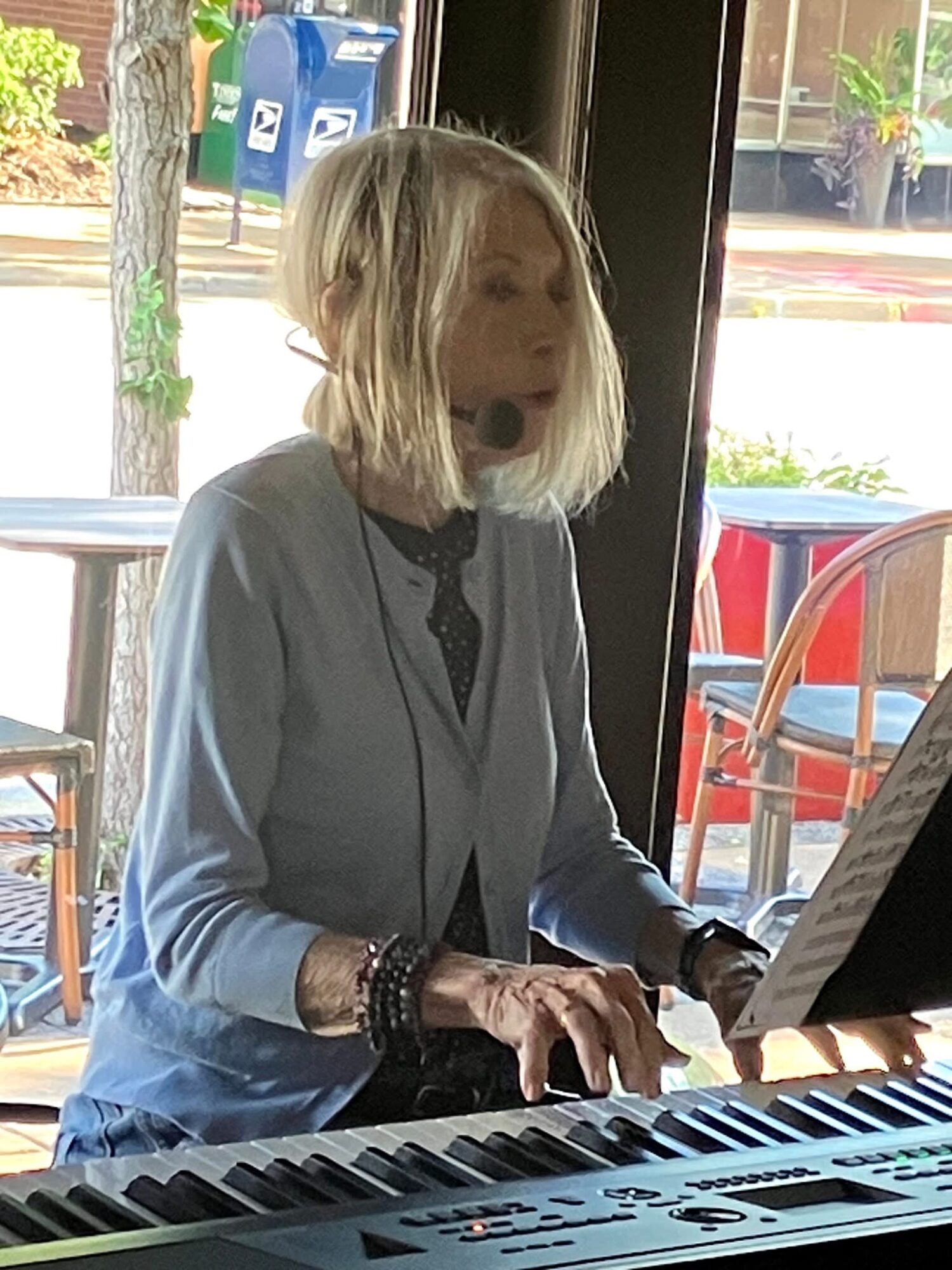 Woman with shoulder-length blonde hair playing a keyboard indoors, sunlight streaming through windows behind her.
