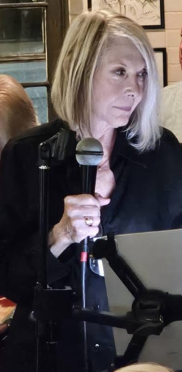 Woman with shoulder-length blonde hair holding a microphone, looking at a laptop screen, in an indoor setting.