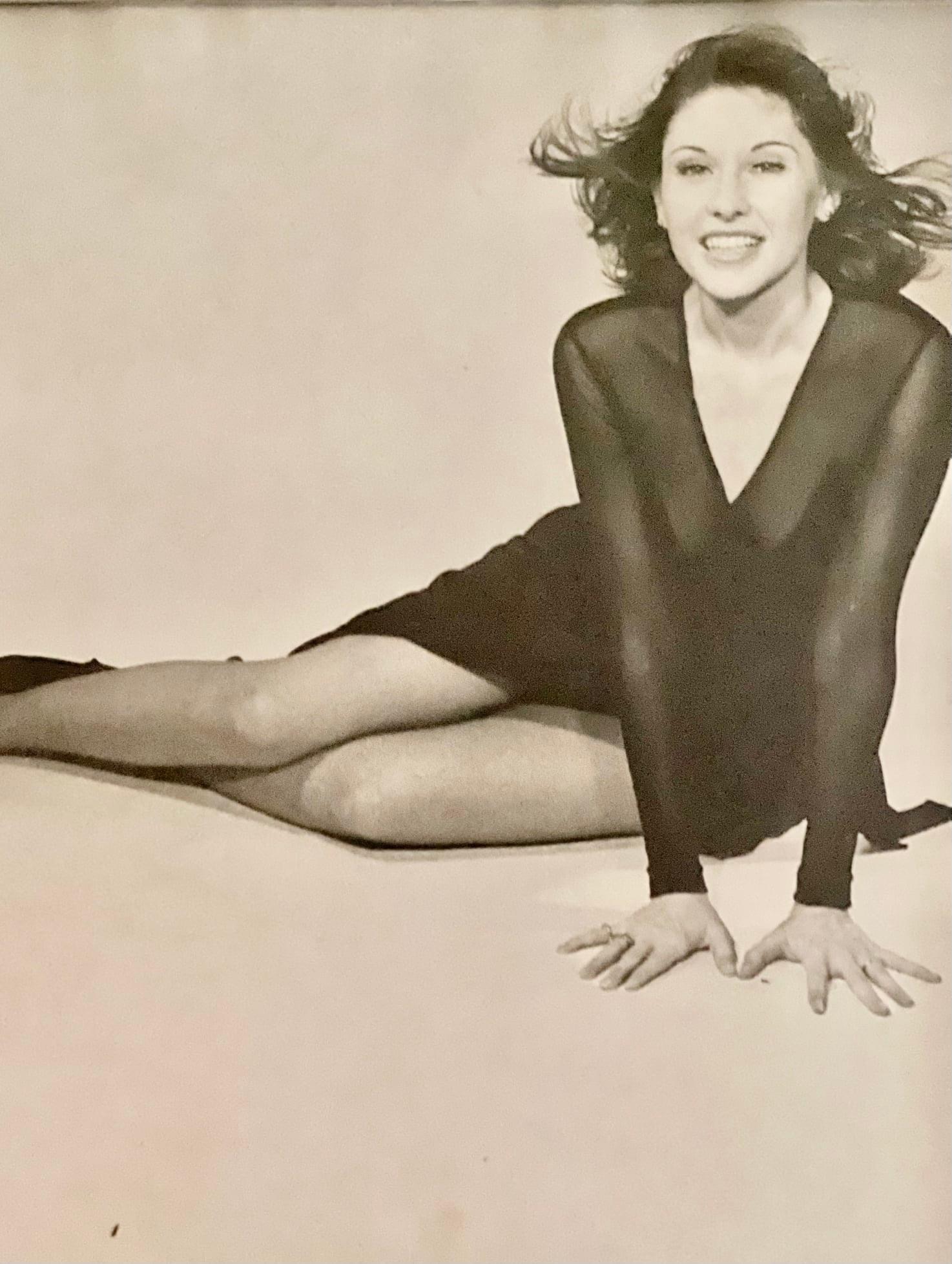 Woman with short wavy hair, smiling, sitting on the floor with legs extended, wearing a dark dress, hands on the ground.