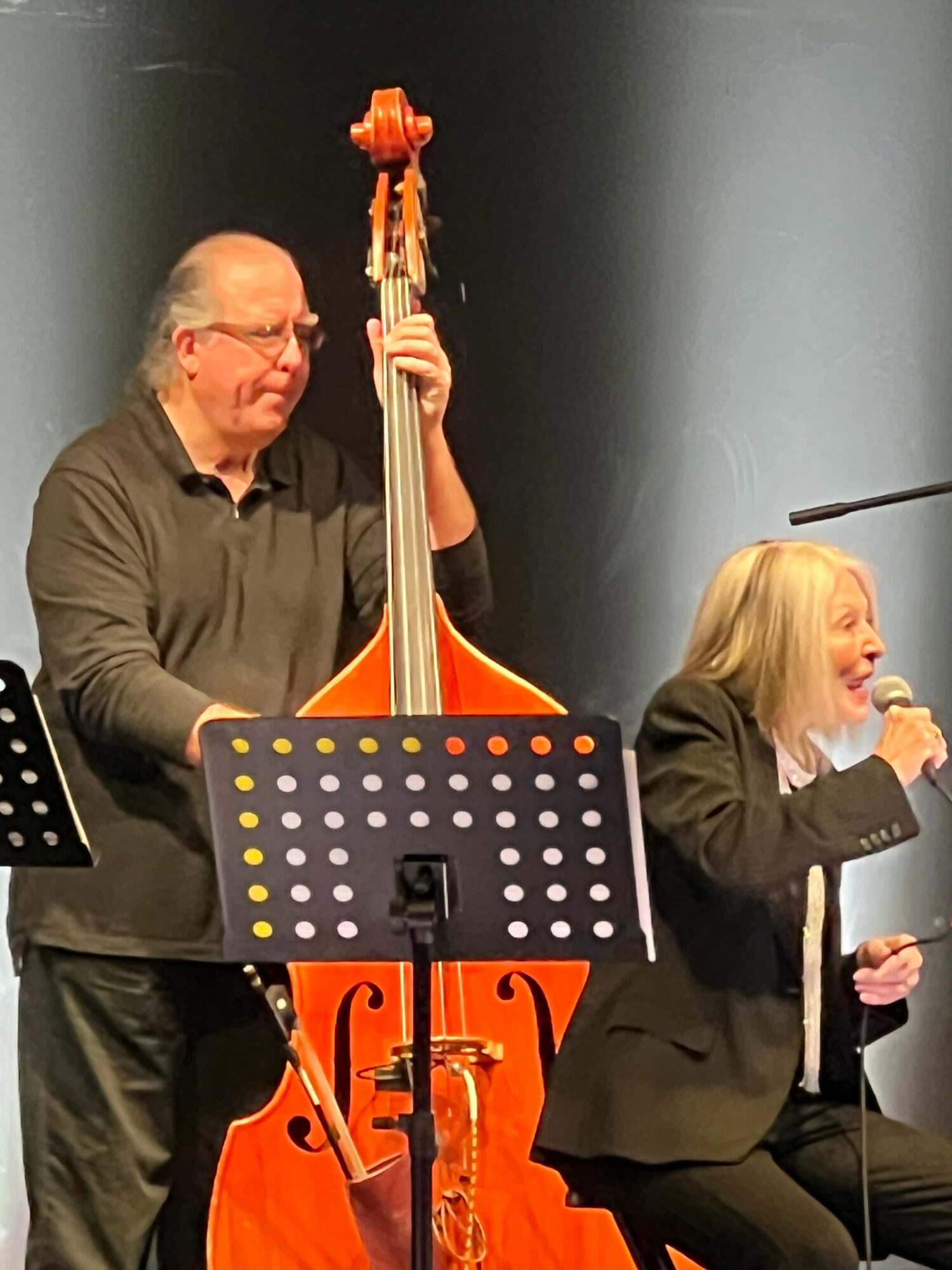 Person playing a double bass, with a music stand in front, and a woman speaking into a microphone in the background.