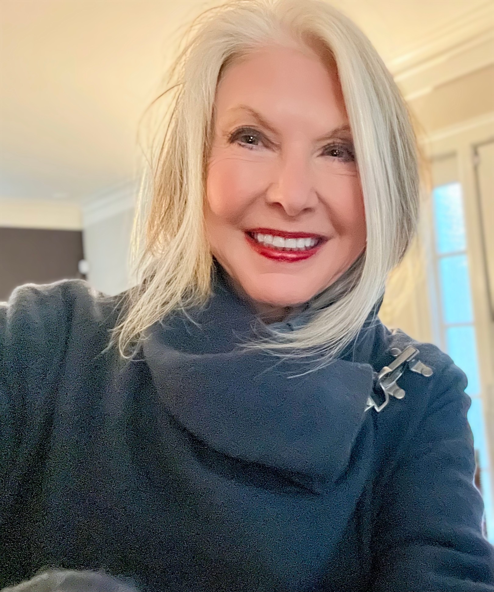 Smiling woman with shoulder-length gray hair, wearing a dark top and scarf, indoors with a window in the background.