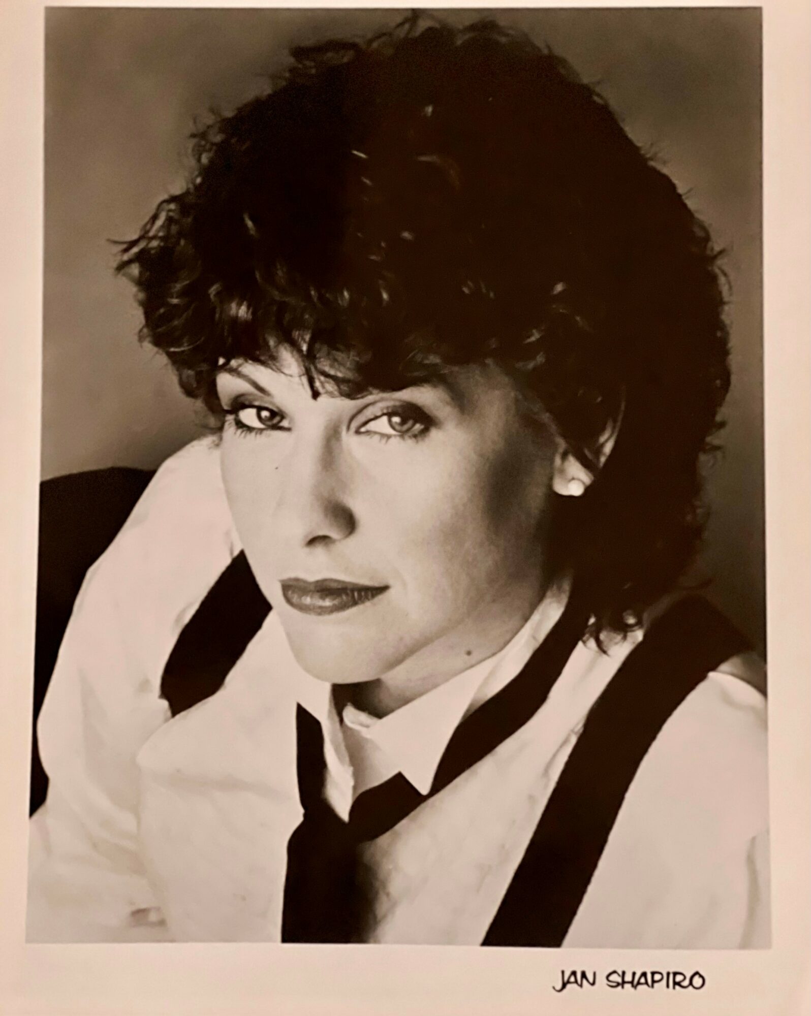 Black and white portrait of a woman with curly hair, wearing a shirt and tie, looking at the camera.
