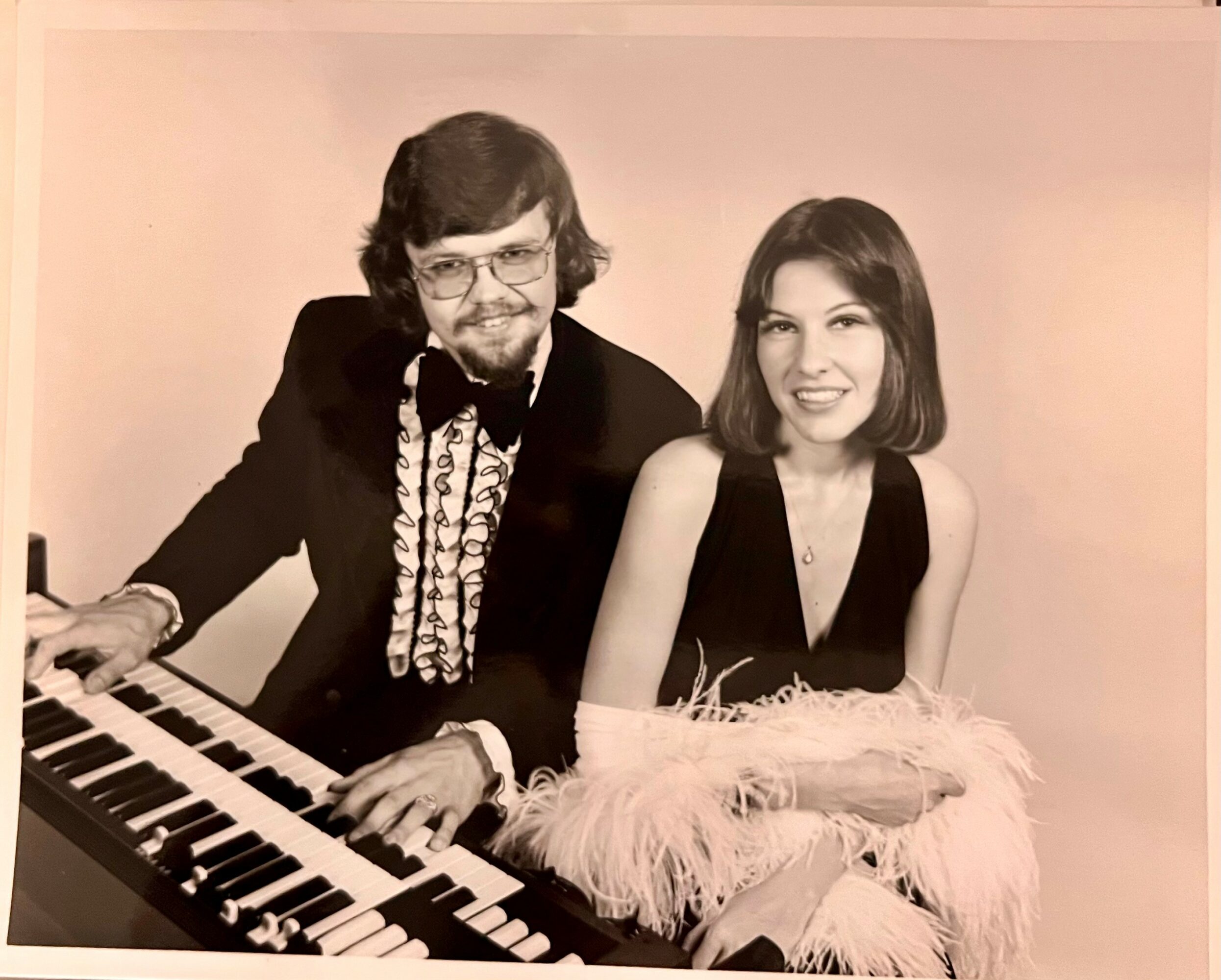A man and woman sit together, with the man playing a keyboard, both smiling, in a vintage photo.