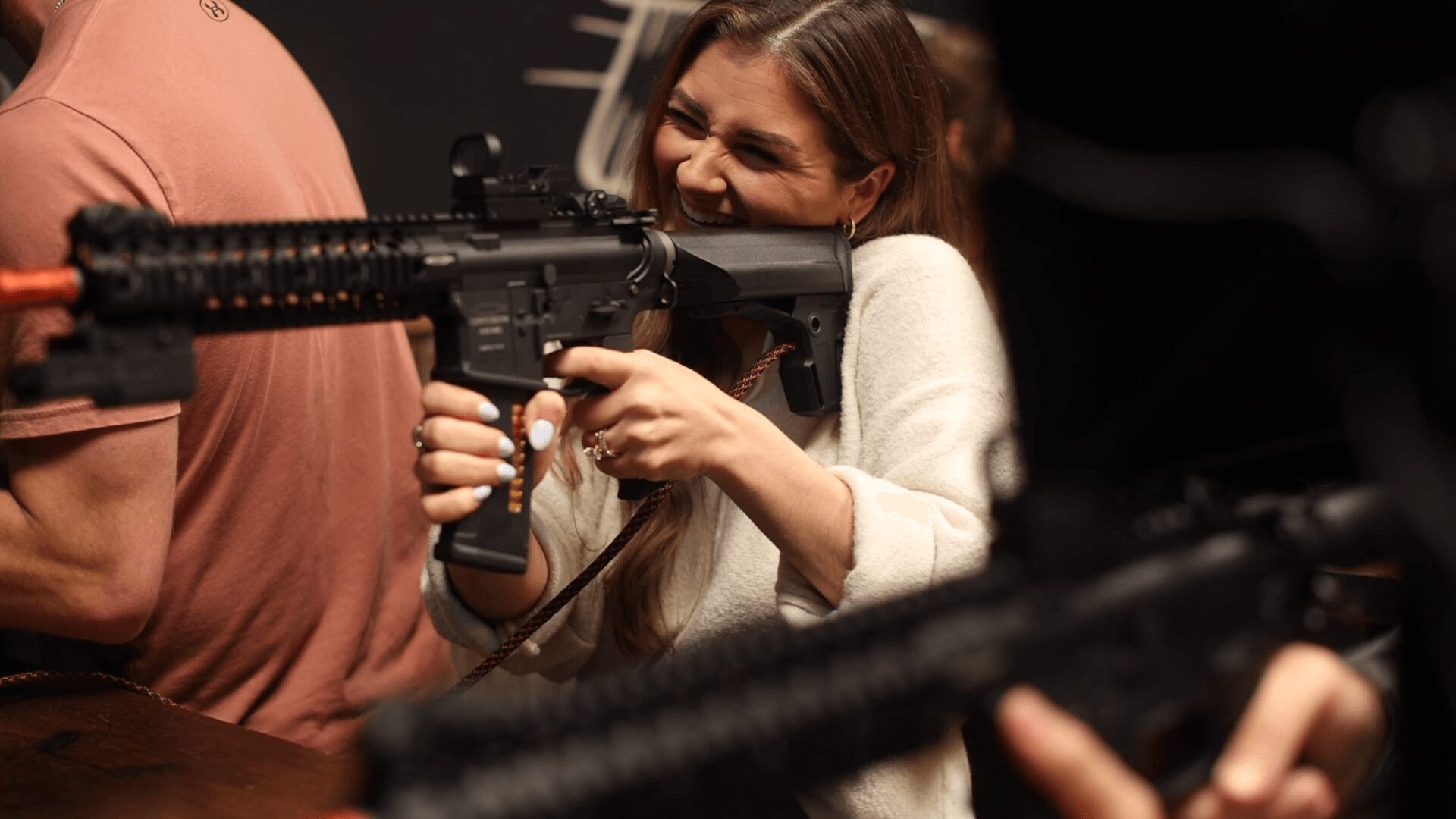 Young girl aiming a toy gun, smiling, with people around her in an indoor setting.
