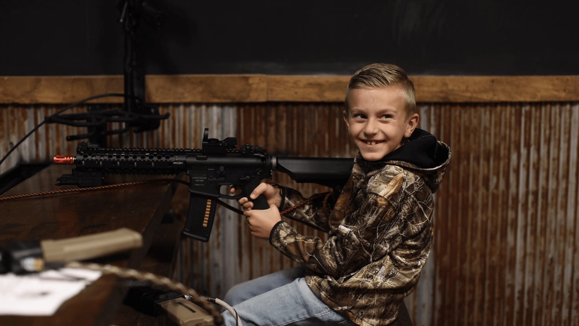Smiling boy in camouflage jacket holding a toy gun indoors, sitting at a table with a wooden wall background.