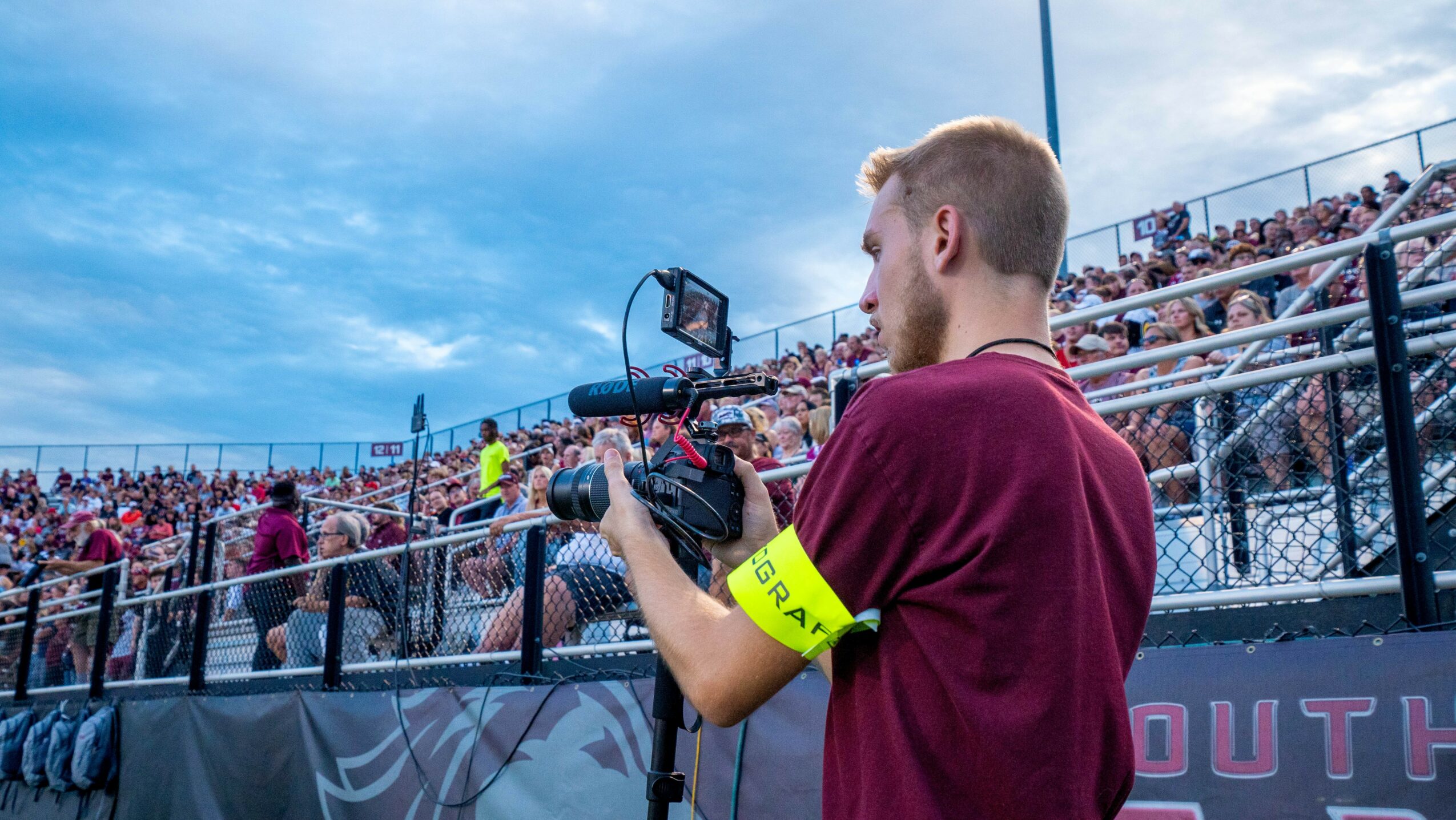 Young man with a camera and yellow armband filming at an outdoor event, crowd in the background, cloudy sky.