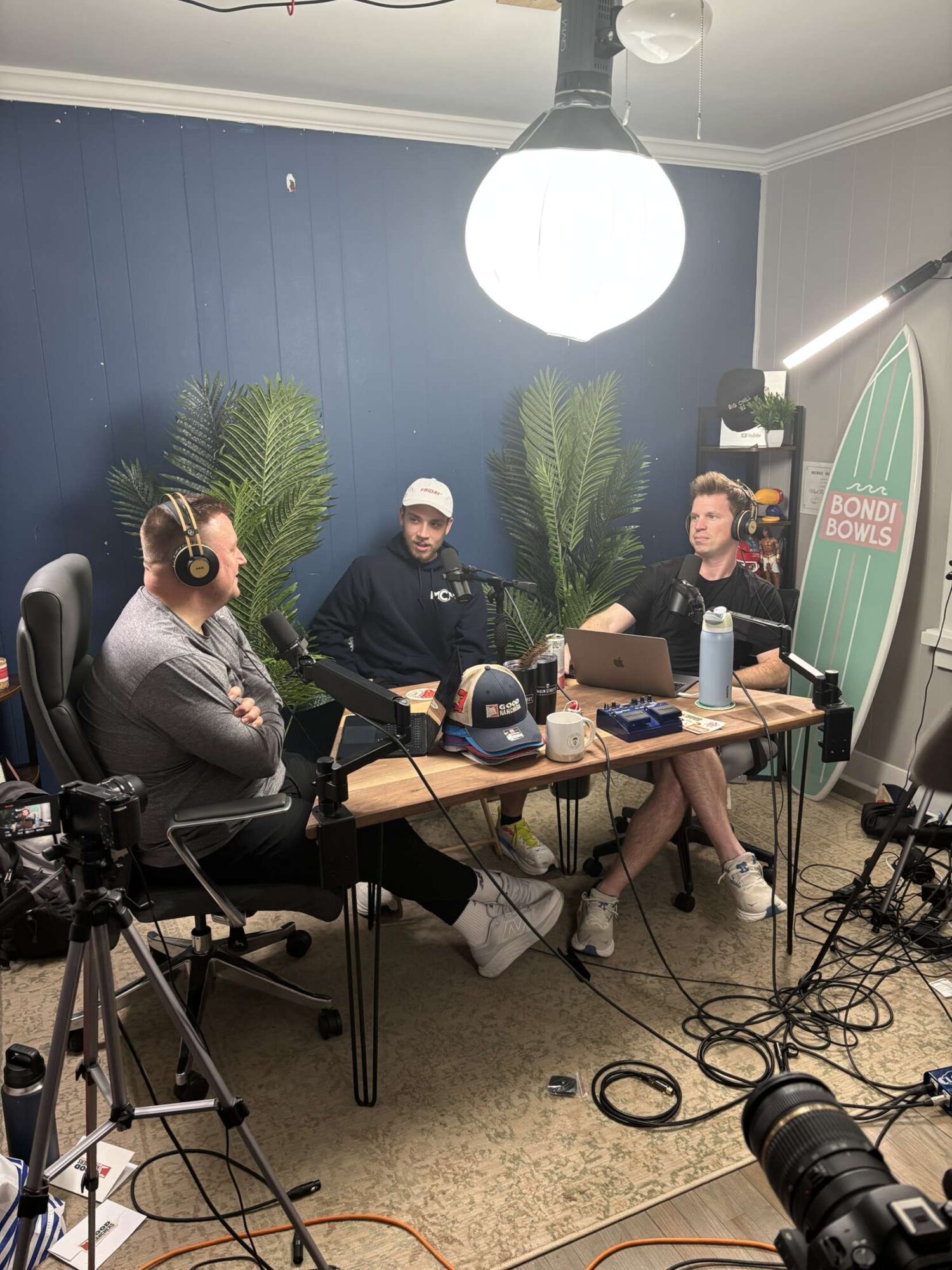 Three people sit at a table with microphones and headphones, recording a podcast or radio show, with a surfboard and plants in the background.