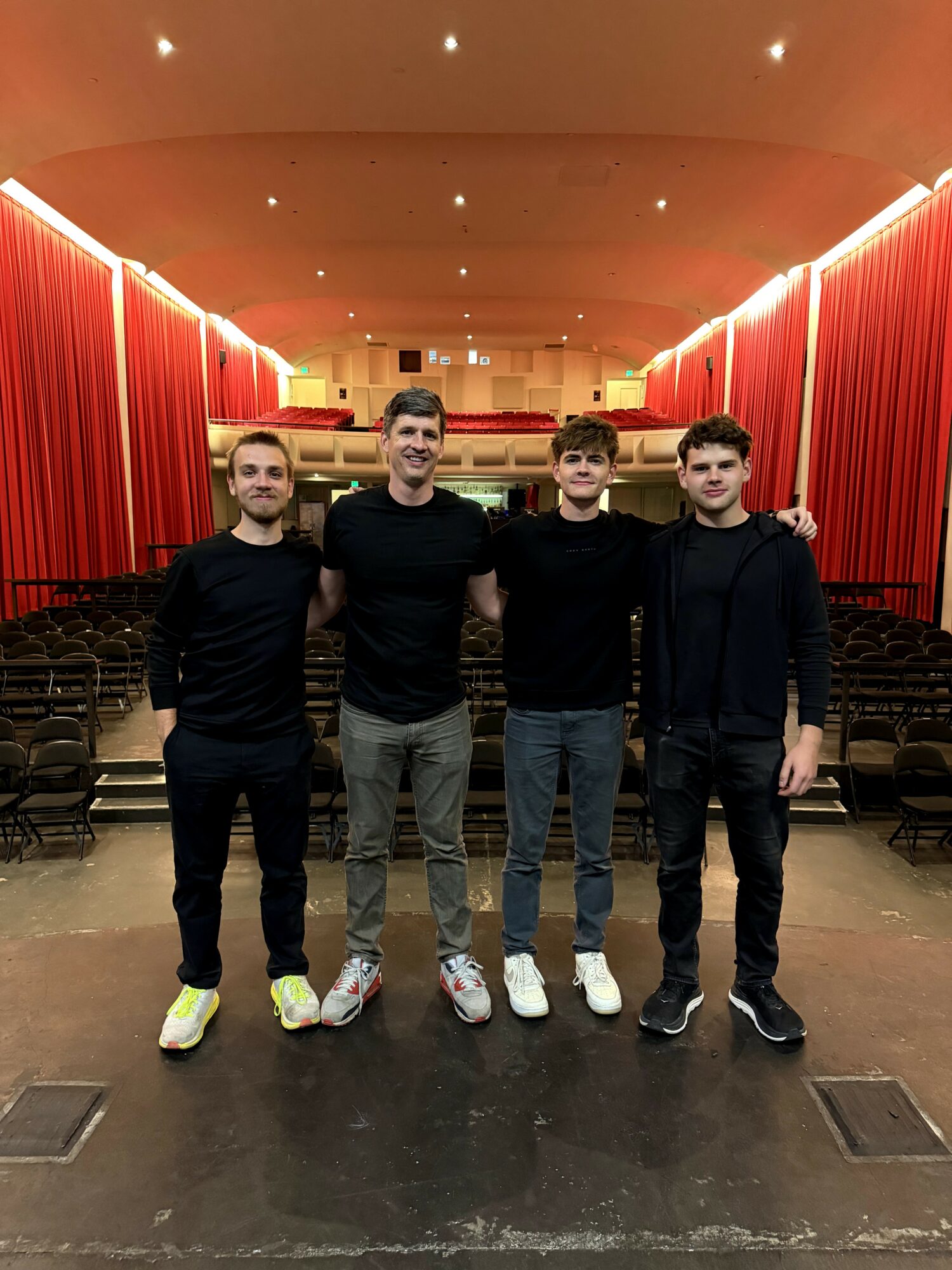Four young men stand on stage in an auditorium with red curtains and seating in the background.