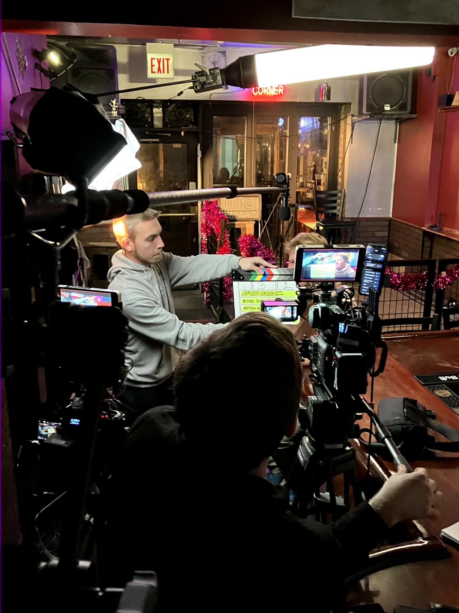 Person in a gray hoodie sitting at a table with filming equipment, in a dimly lit indoor setting with pink walls.