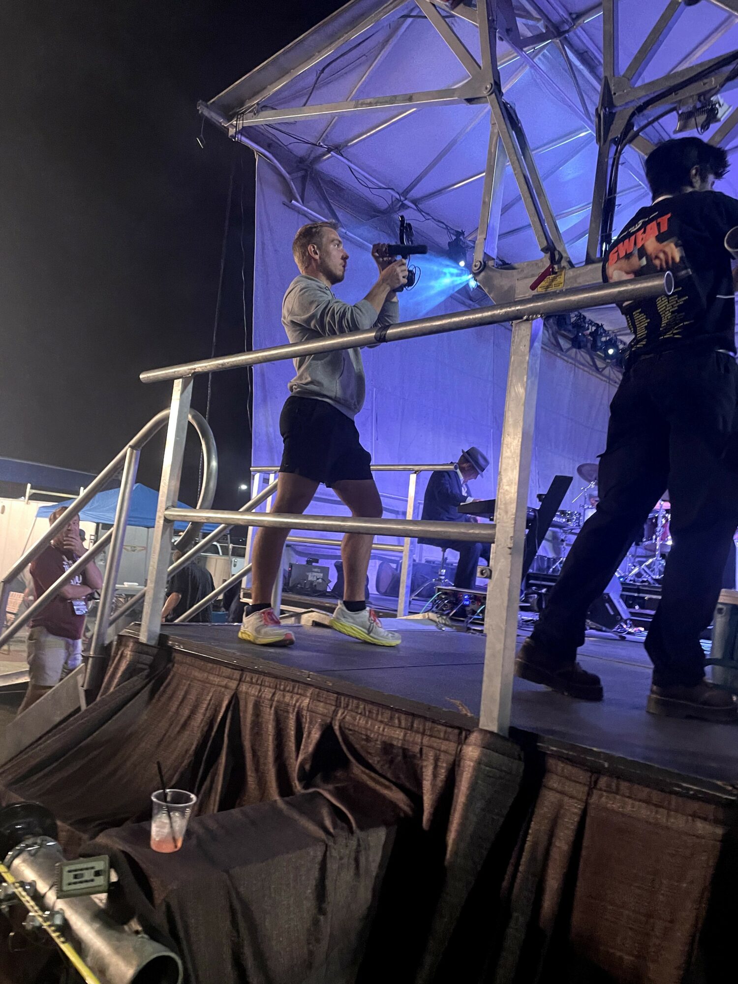 Person on stage wearing shorts and a mask, holding a microphone, with another person nearby, stage setup visible.