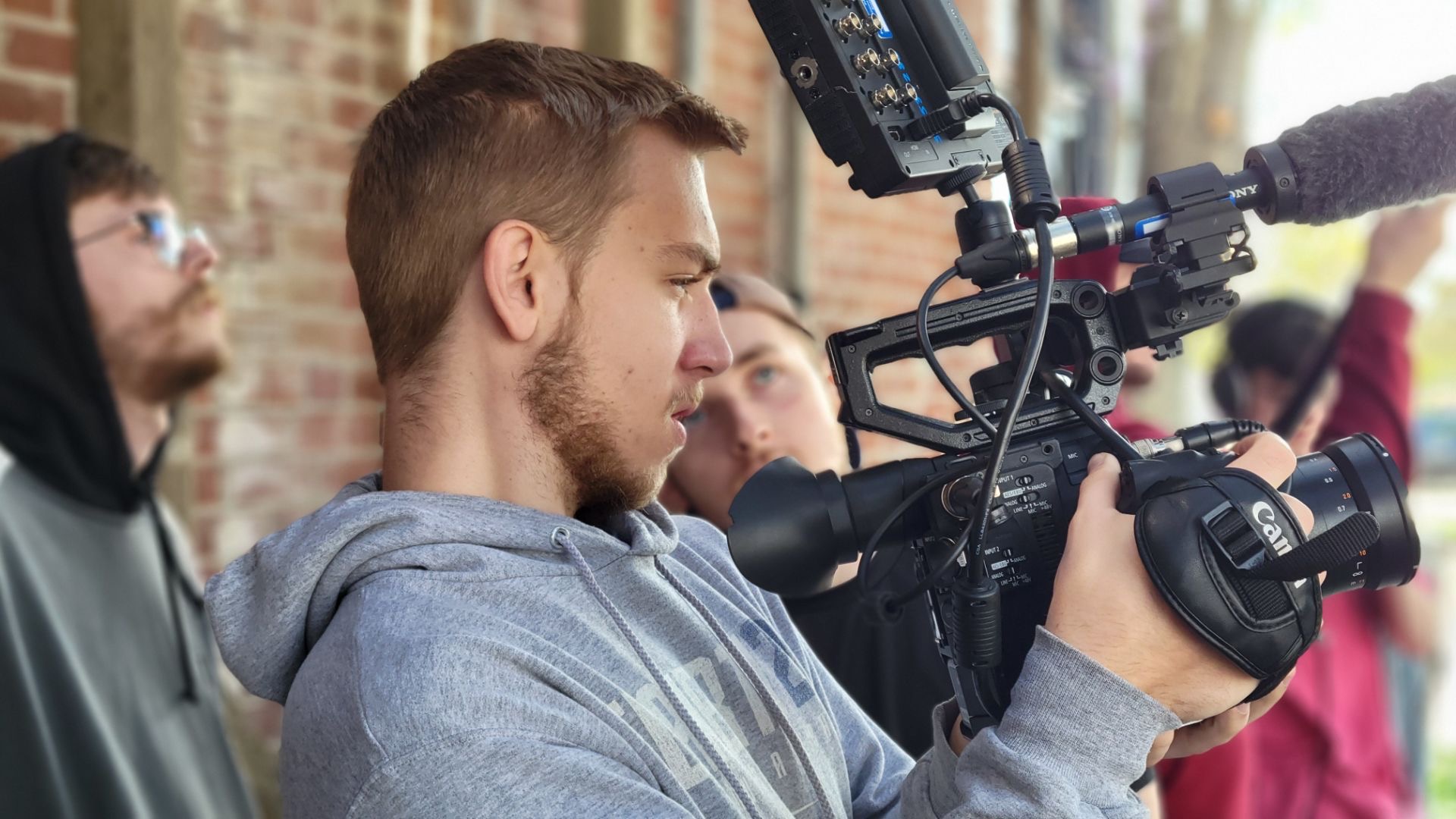 Person operating a professional video camera outdoors with others in the background.