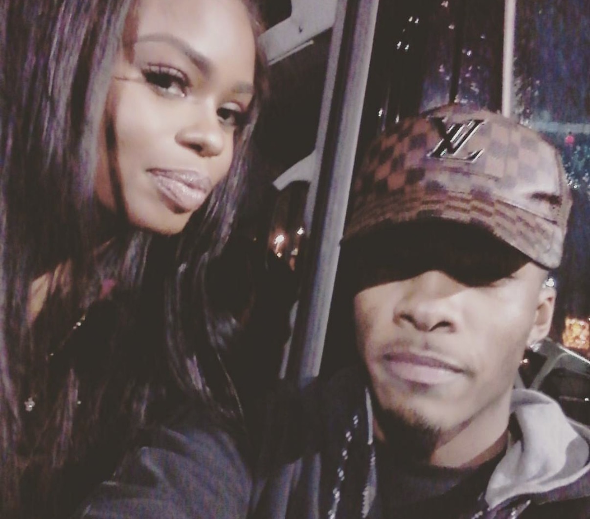 Two people taking a selfie inside a vehicle at night, one wearing a cap and the other with long hair, smiling.