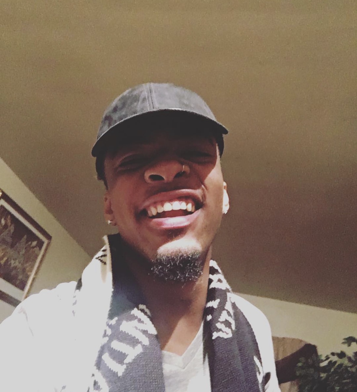 Person smiling, wearing a cap and scarf, indoors with a ceiling and wall in the background.