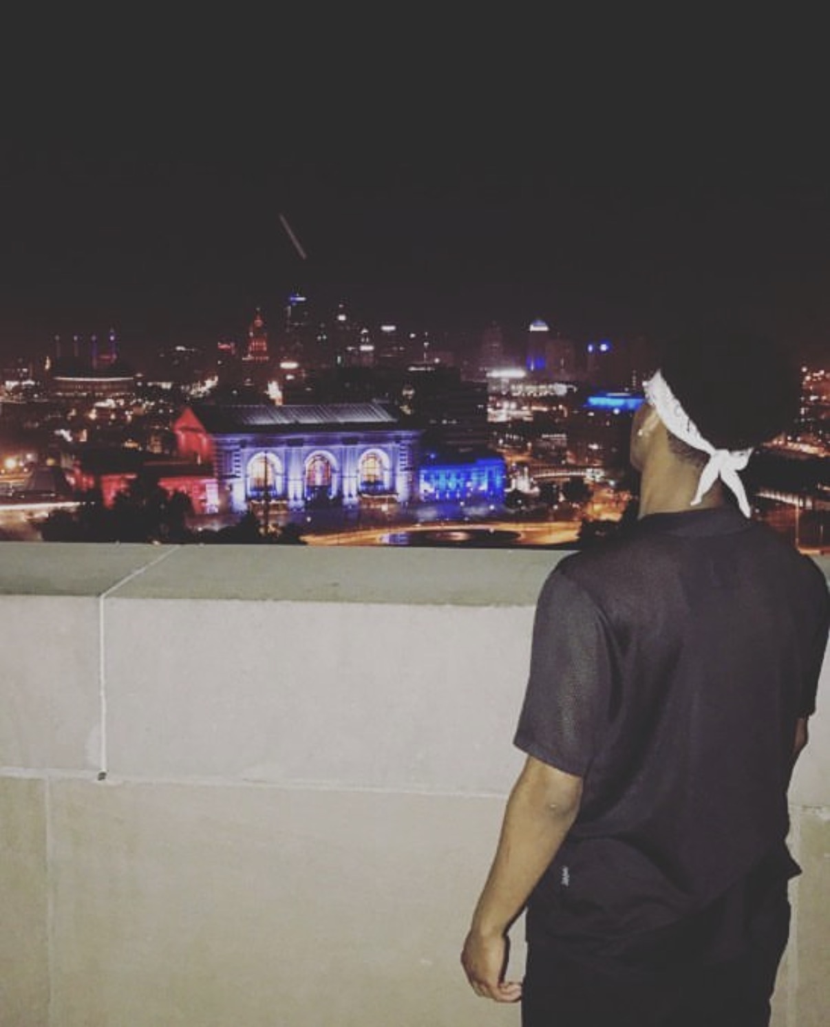 Person with a white headband looking at a city skyline at night, illuminated buildings and lights in the background.