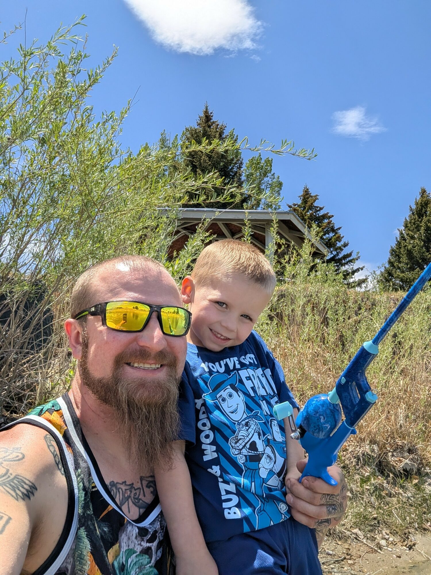 Man with sunglasses and a young boy outdoors, holding a blue toy gun, surrounded by trees and blue sky.