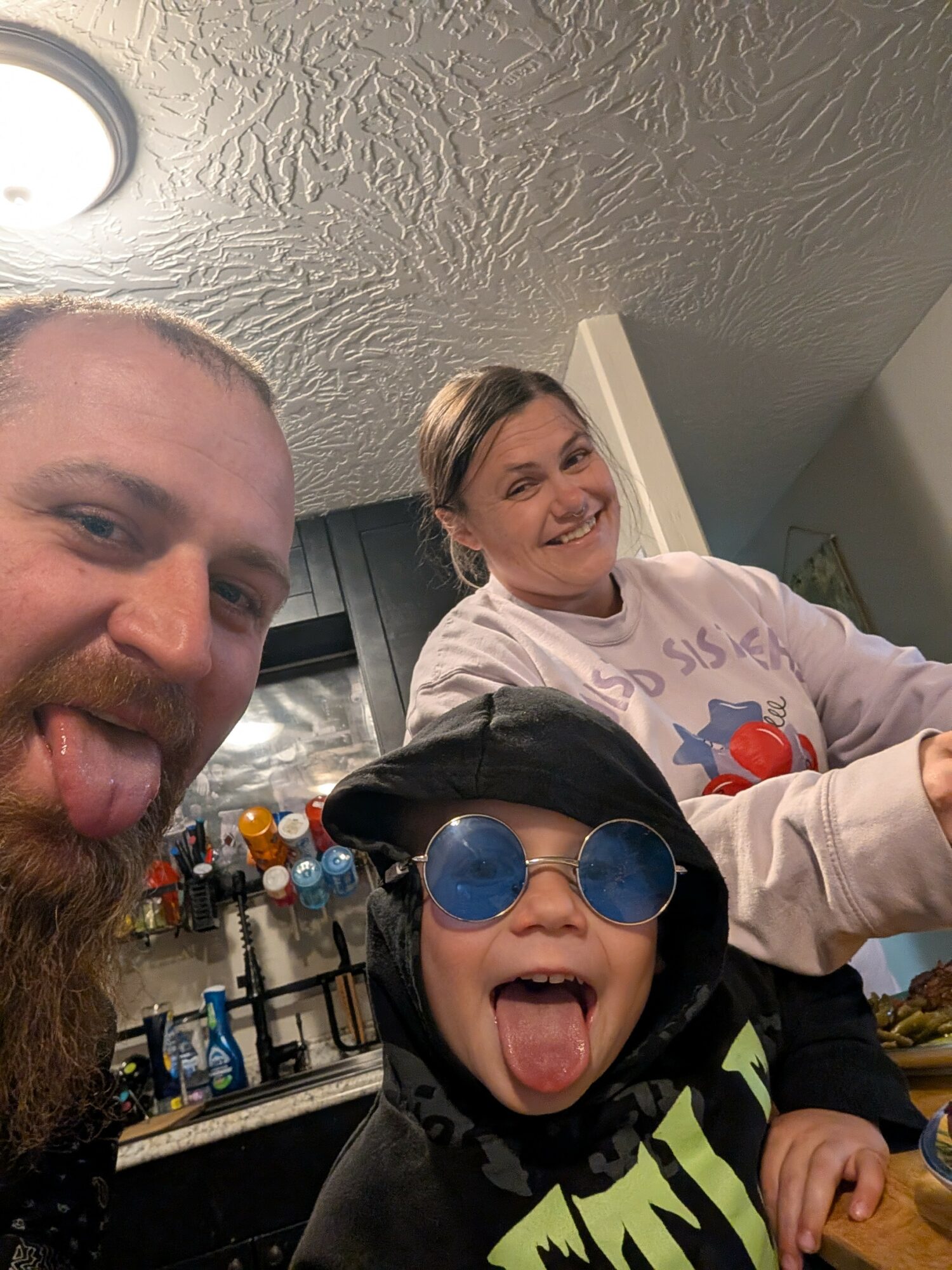 Three people smiling and sticking out their tongues in a kitchen, with a textured ceiling and bottles in the background.