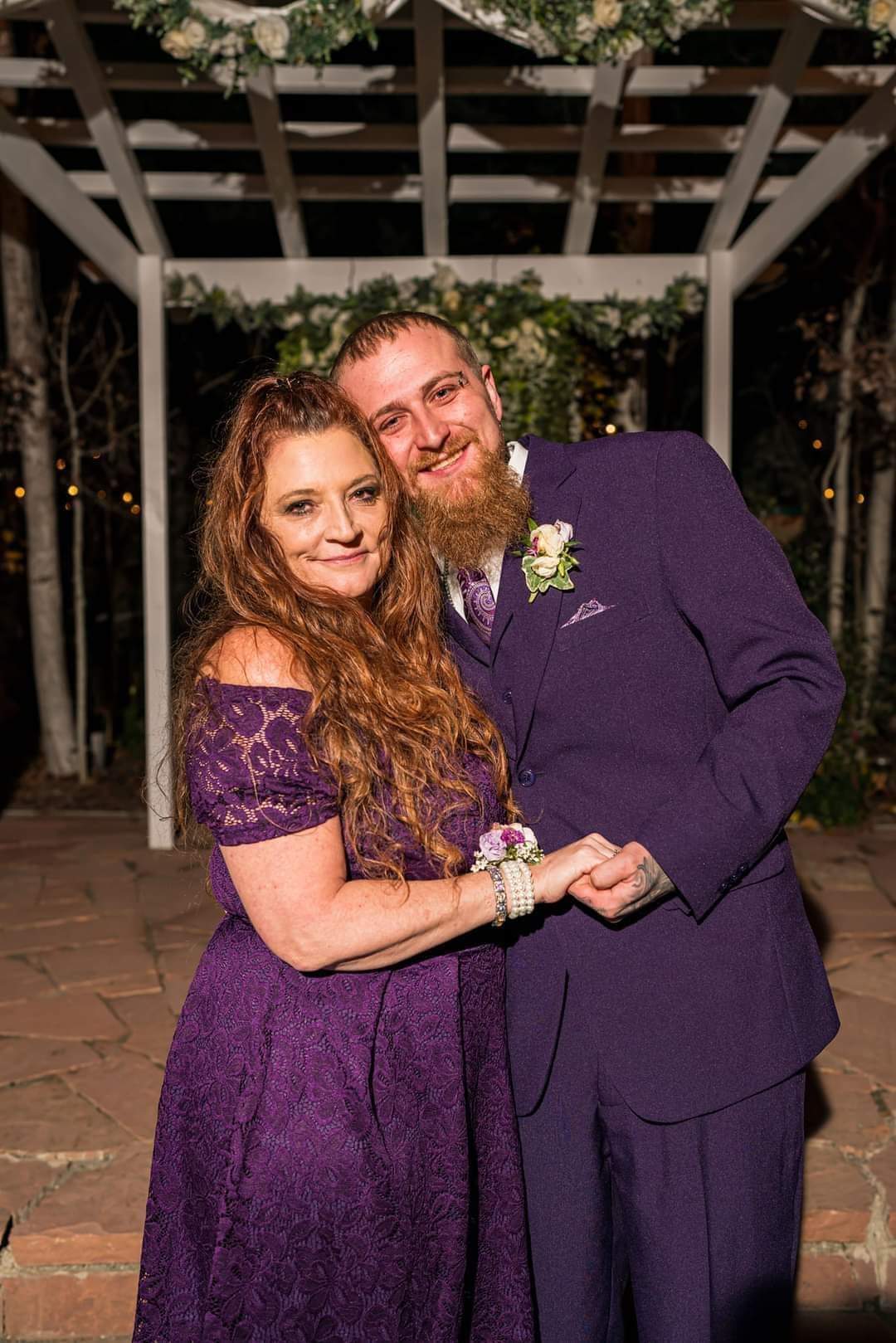 A smiling couple, woman with long wavy hair in a purple dress and man with a beard in a dark suit, pose outdoors at night.