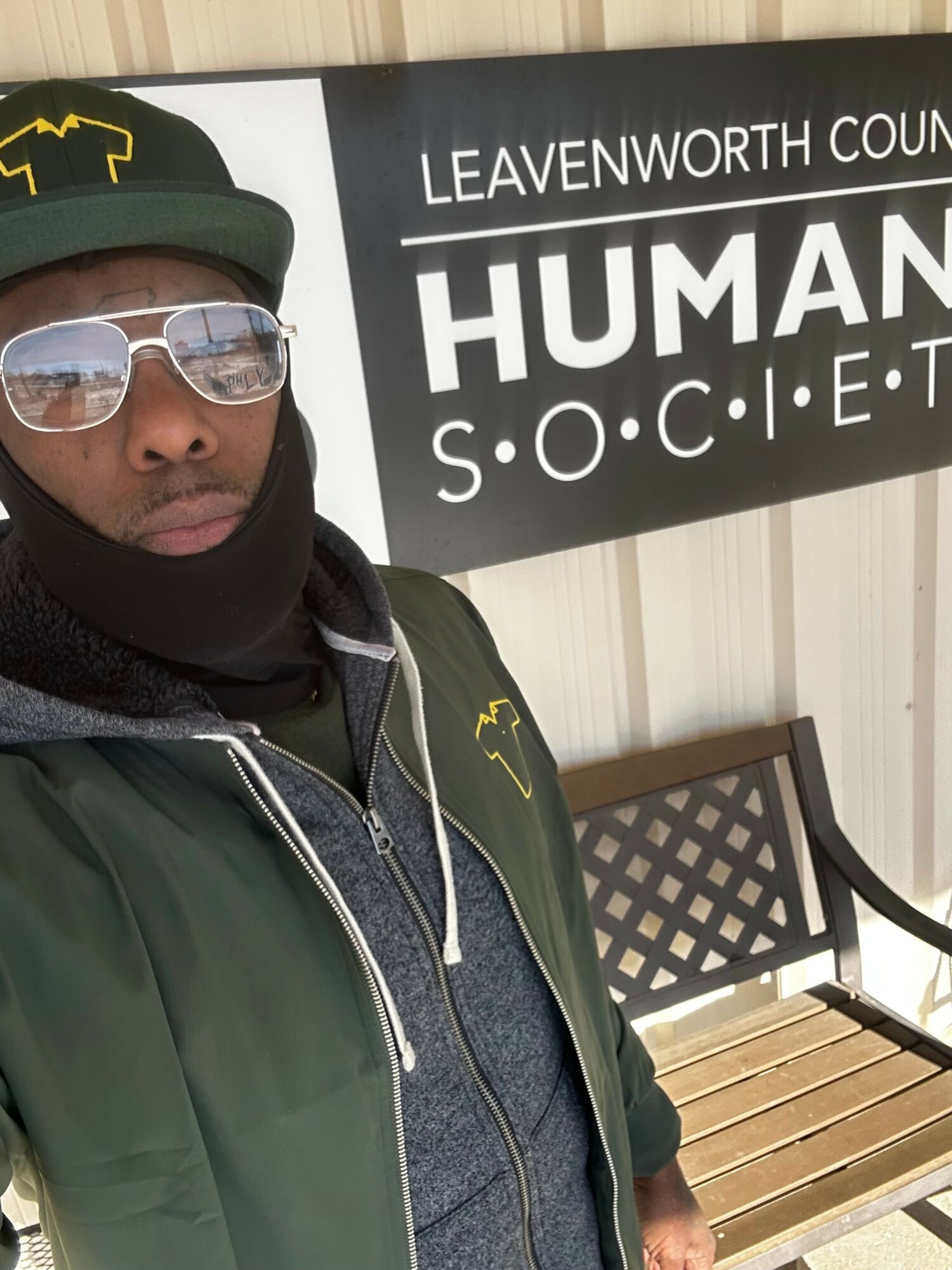 Person wearing sunglasses and a cap standing outdoors near a sign that reads 'LEAVENWORTH COUNTY HUMAN SOCIETY'.