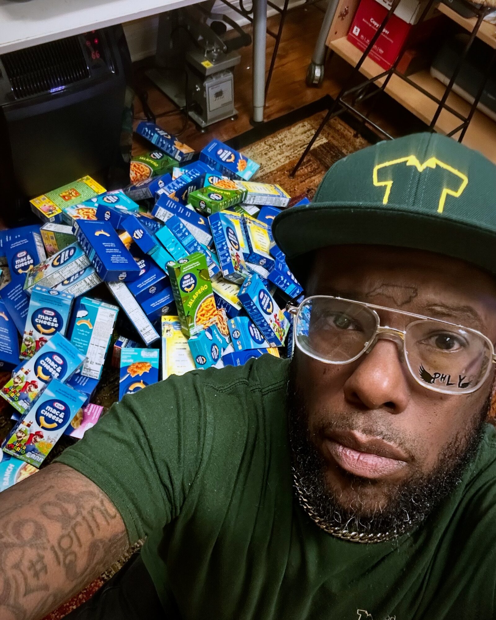 Man wearing glasses and a cap with a yellow logo takes a selfie in front of a large pile of blue and green snack packages.