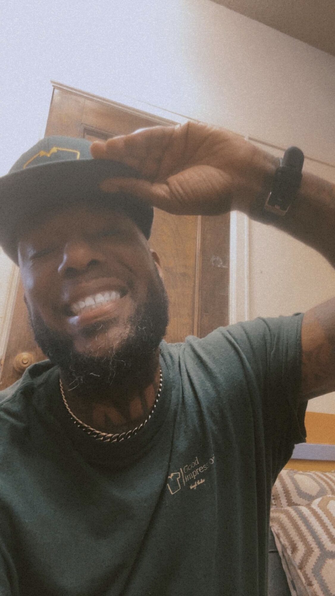 Person smiling, wearing a cap and a dark green shirt, saluting with hand on cap, indoors.
