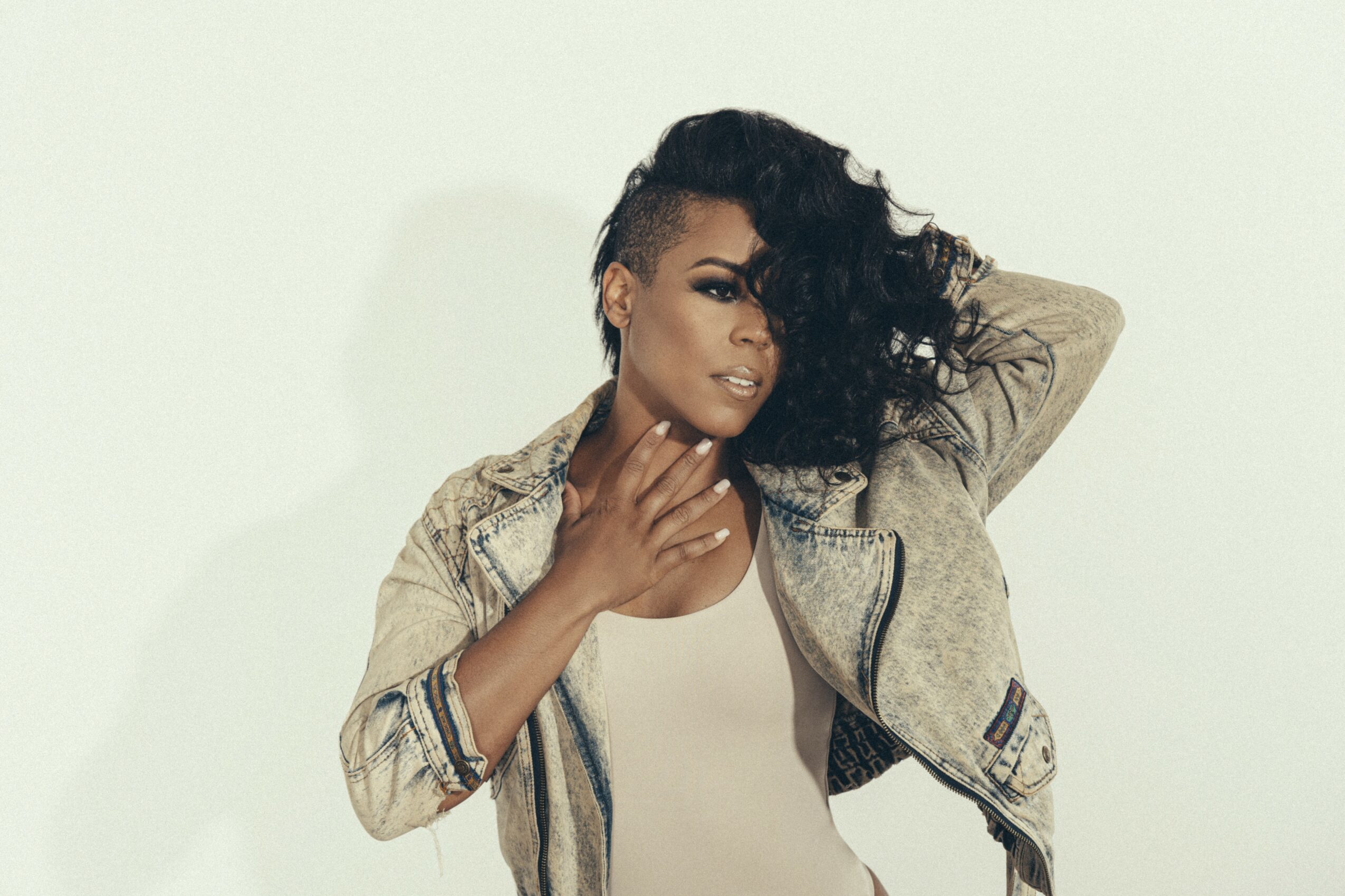 Woman with curly hair and makeup, wearing a beige jacket and white top, posing with hand near neck against a plain background.