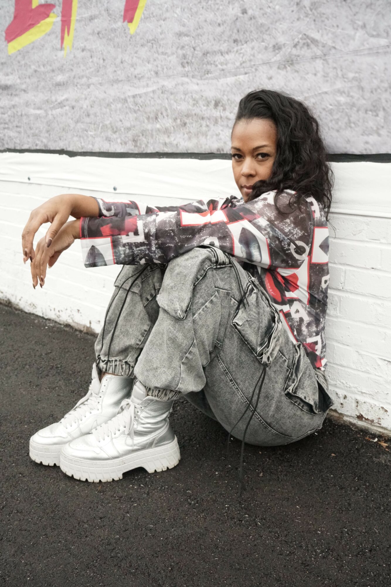 Person sitting against a wall, wearing a camouflage jacket, jeans, and white boots, with a skateboard nearby.