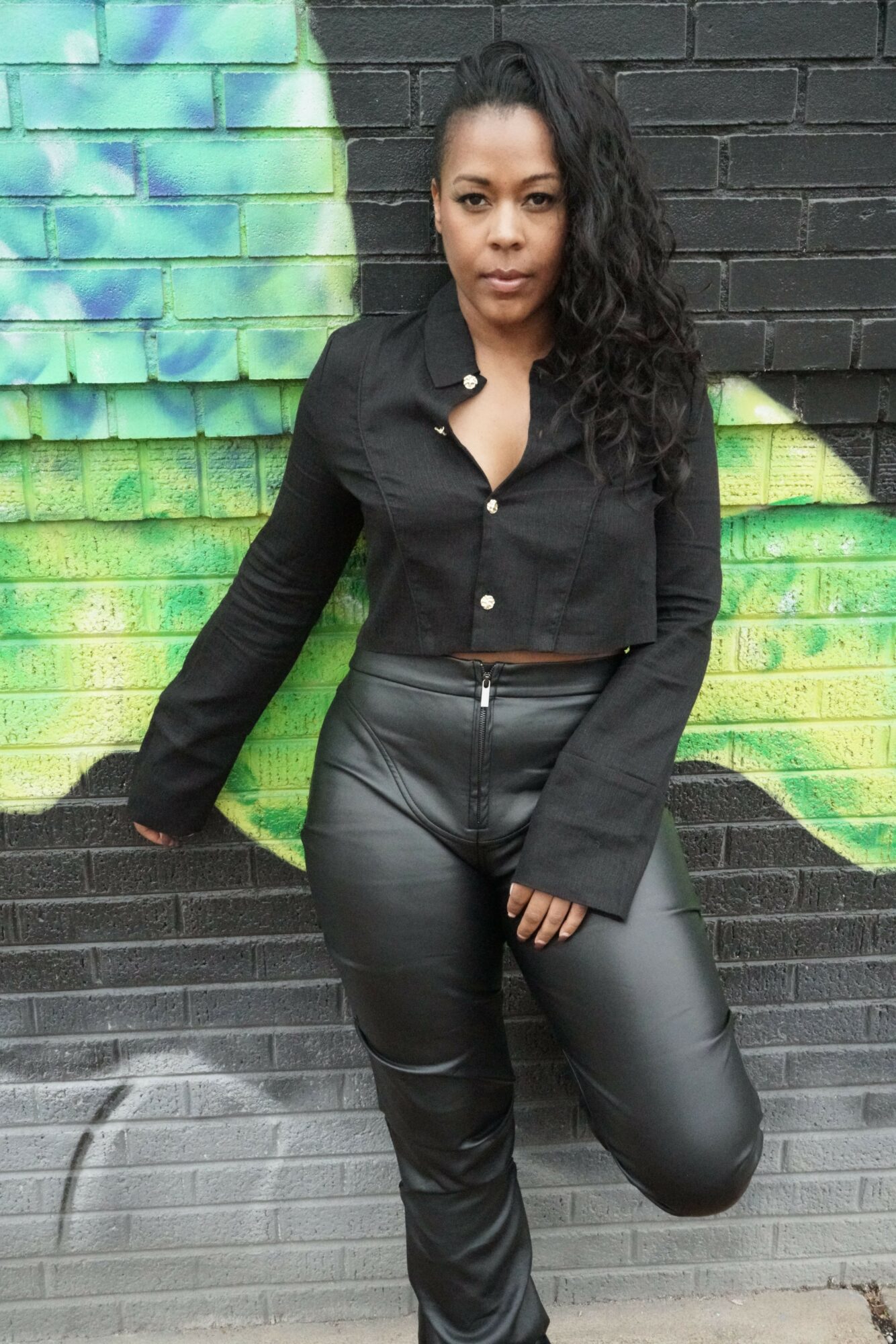 Woman lying on a colorful blanket on a dark brick surface, wearing black leather pants and a black top, looking at the camera.