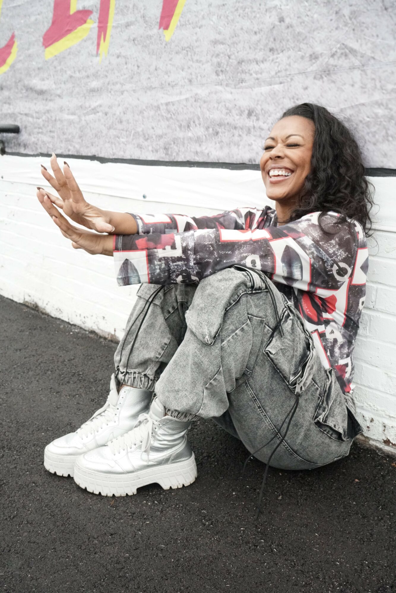 Person lying on the ground with legs crossed, smiling, wearing white boots, jeans, and a patterned top, against a wall.