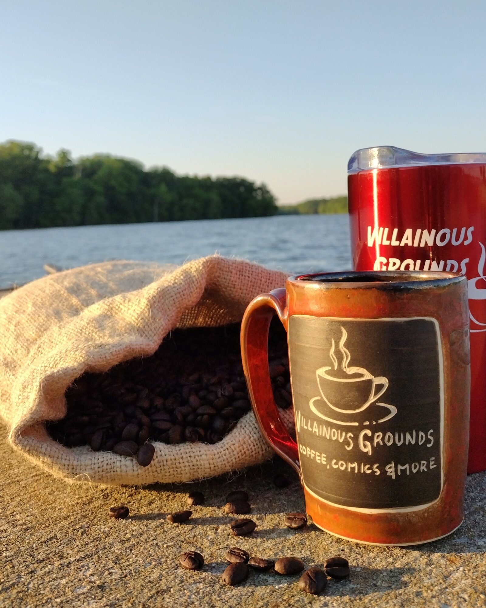 Coffee beans spilling from a beige bag, two mugs with logos, outdoors near a body of water, trees in background.