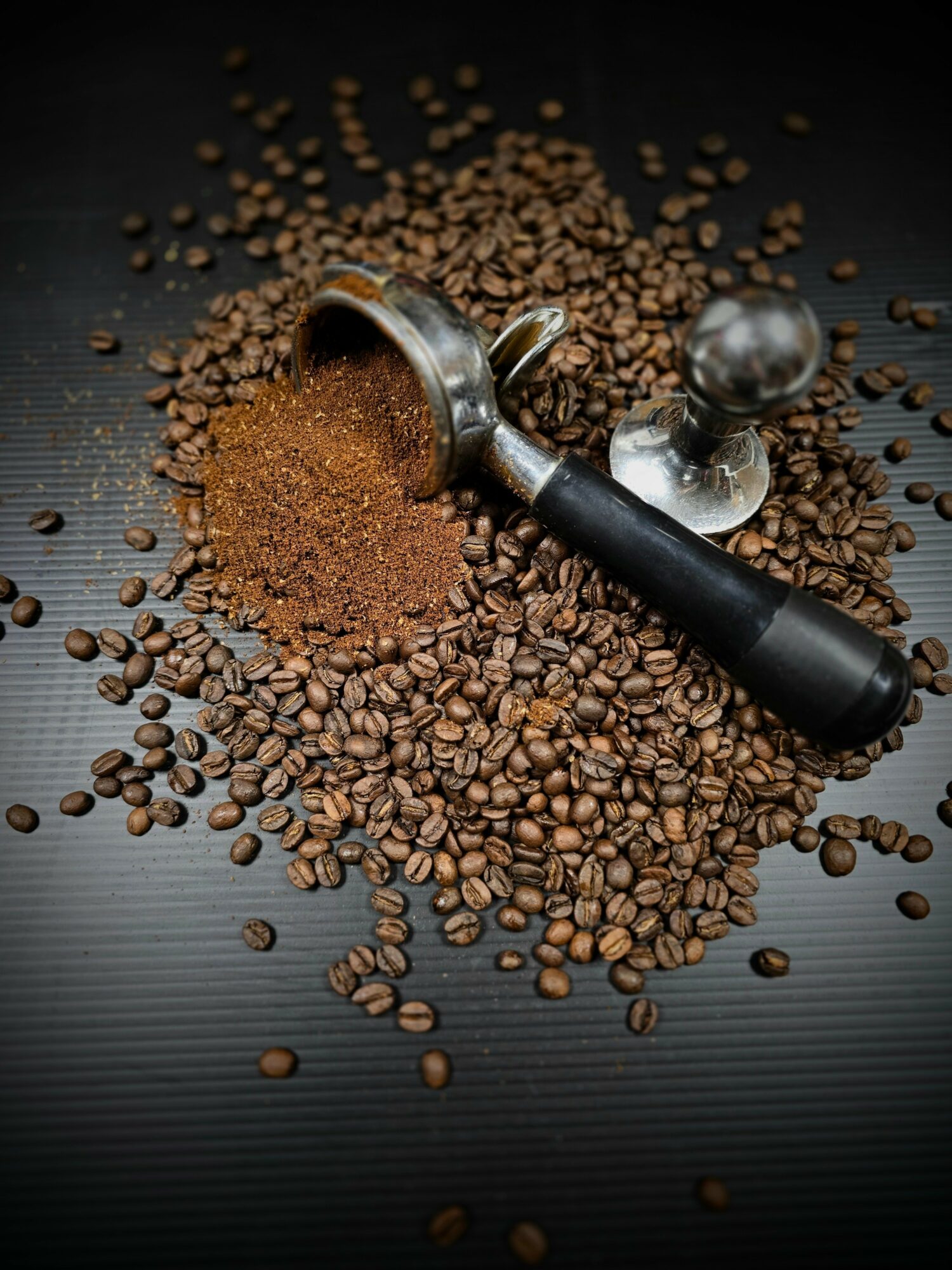 Coffee beans scattered with a coffee scoop and a tamper on a dark surface.