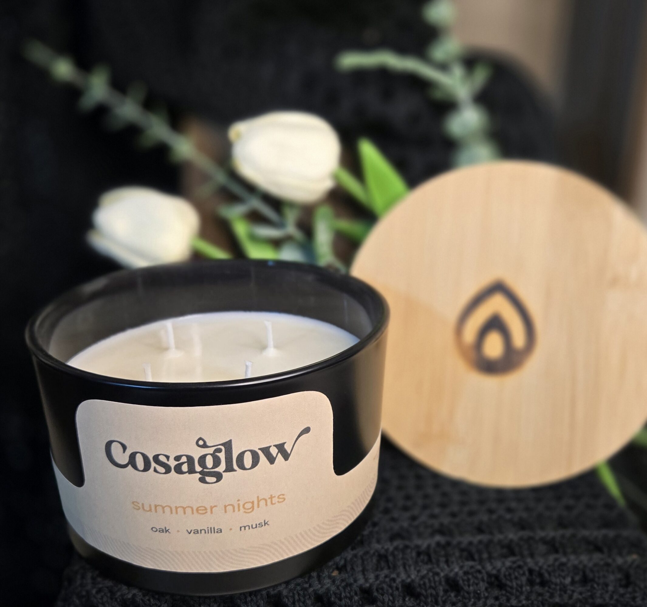 A black candle with a label reading 'Cosaglów summer nights' and a wooden lid with a logo, with white flowers in the background.