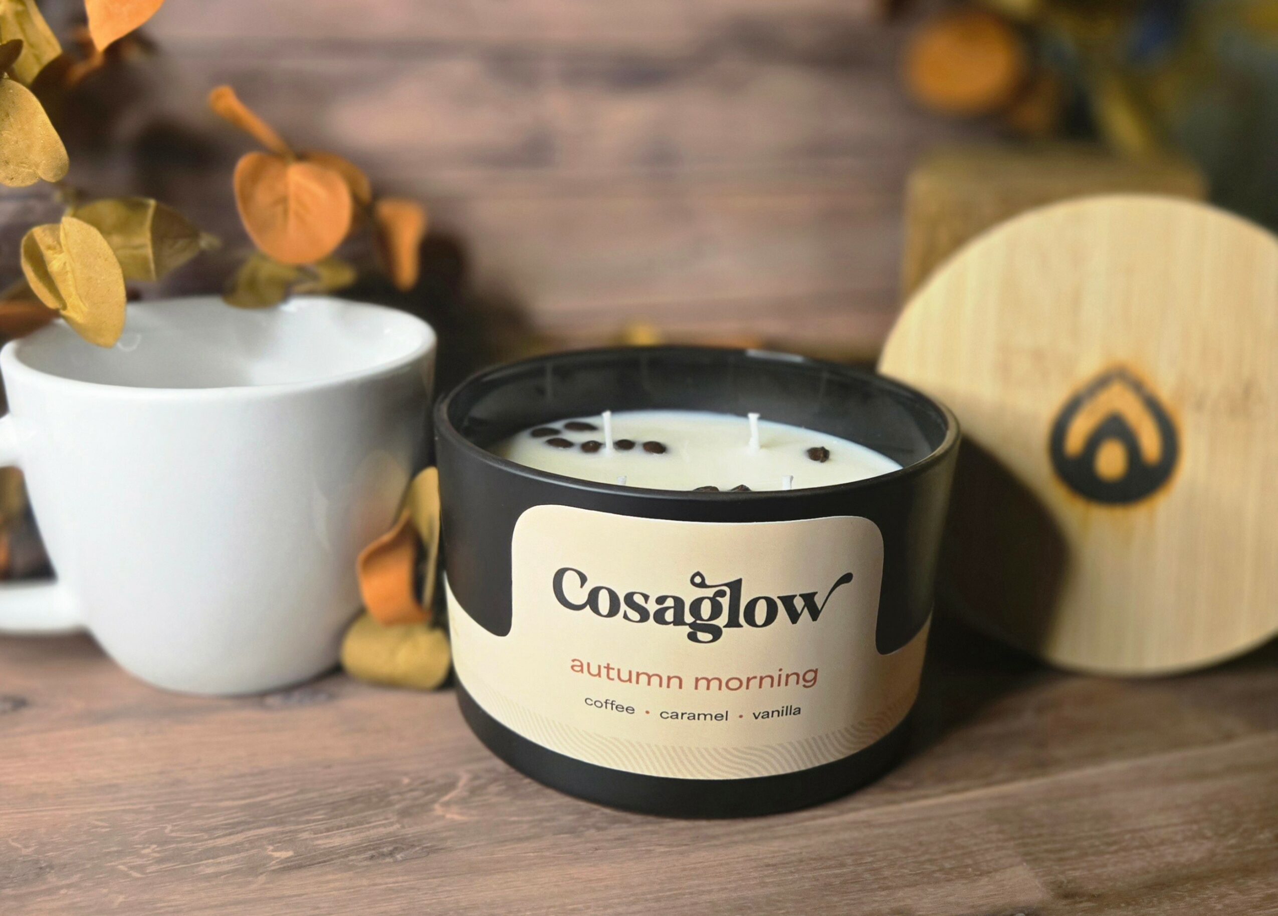 A black candle labeled 'Cosaglow autumn morning' on a wooden surface, with a white cup and a wooden object nearby.