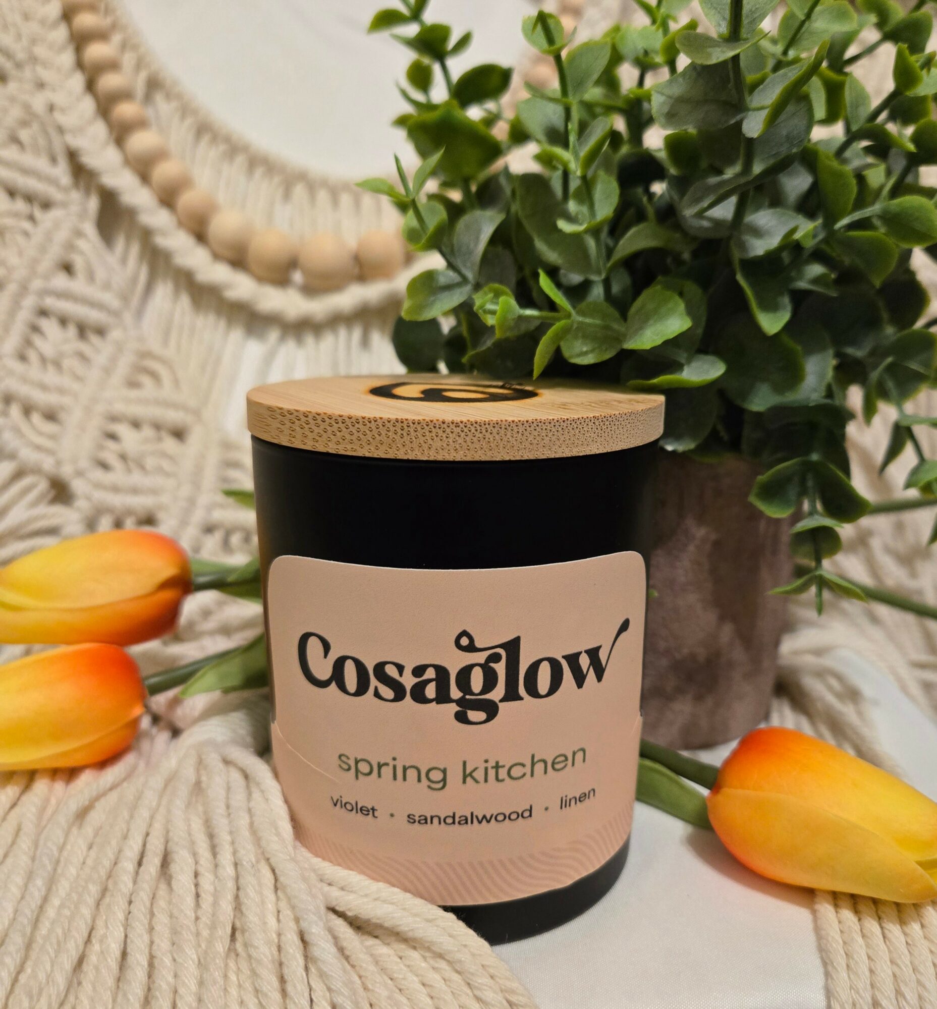 A black candle with a wooden lid, labeled 'Cosaglow spring kitchen,' next to a potted plant and tulips on a textured surface.