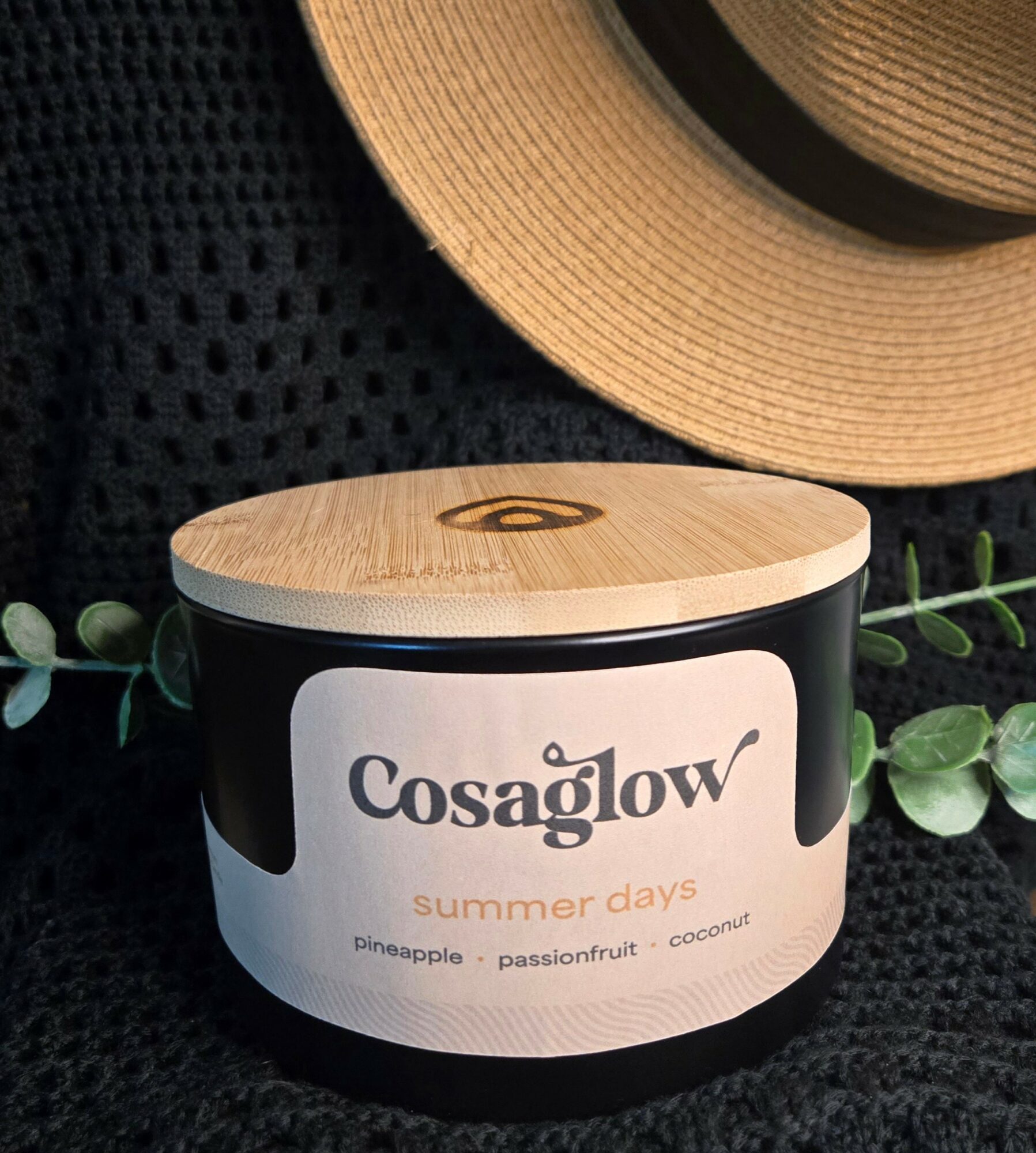 A black container with a wooden lid, labeled 'Cosaglow summer days,' placed on a black textured surface with a straw hat nearby.