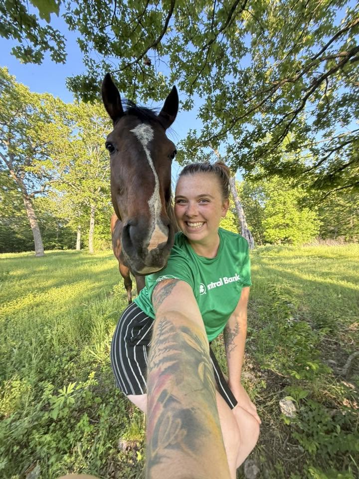 Young woman smiling with a horse outdoors in a green park, trees and blue sky in background.