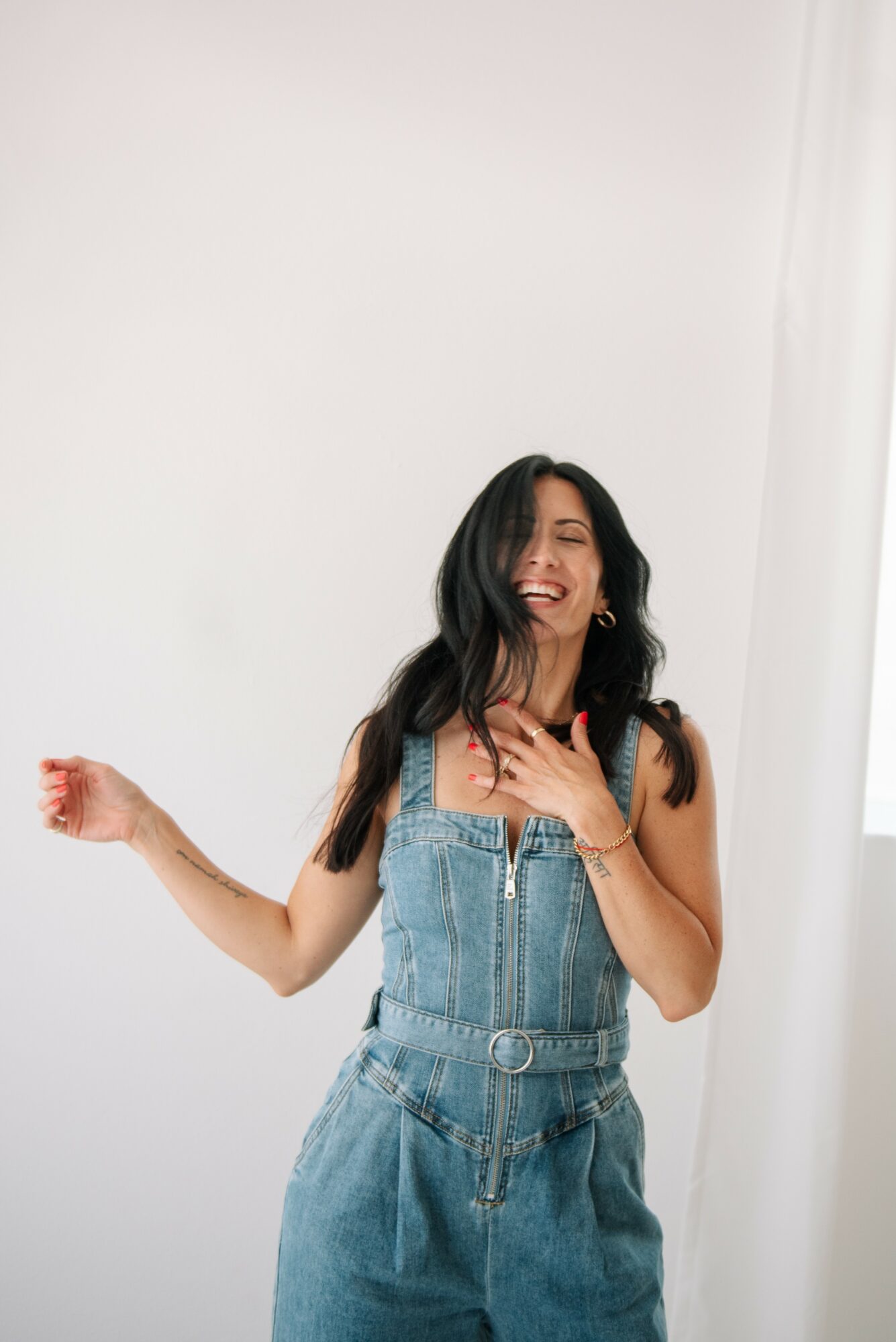 Woman with long dark hair laughing, wearing denim overalls, standing against a plain white wall.