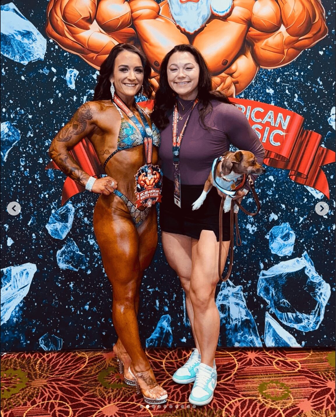 Two women smiling, one in a colorful bikini with a muscular physique, the other in a purple top and black skirt, holding a dog, standing in front of a muscular cartoon backdrop with ice and a red banner.