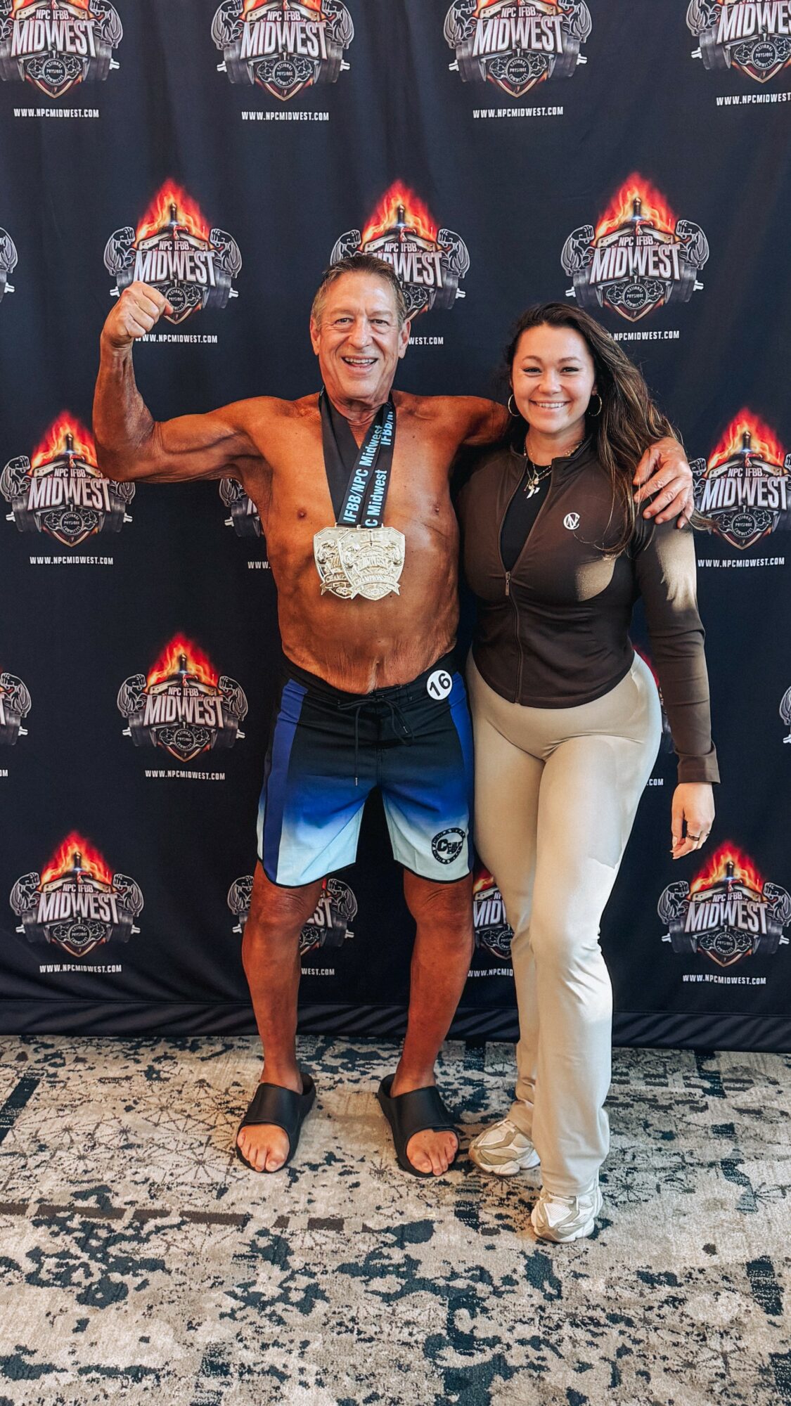 Man and woman standing together, smiling, with a backdrop featuring a logo, man flexing his arm, woman with arm around him.